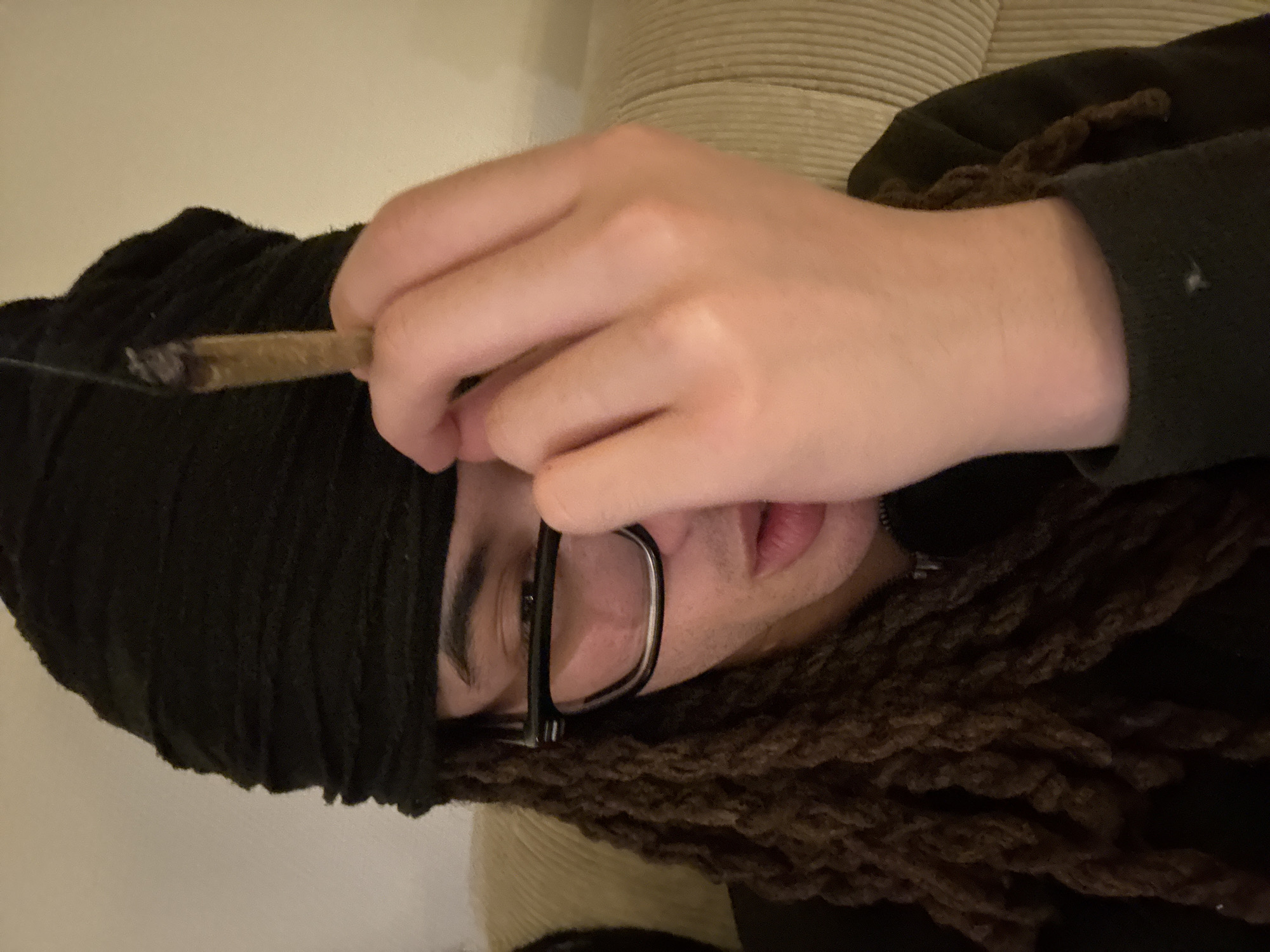 A person is depicted in a close-up, horizontally oriented shot, appearing to be indoors. The individual has light skin and is wearing a dark beanie or head covering, as well as dark-framed glasses. Brown dreadlocks are visible, framing their face and extending downwards. They are dressed in a dark hooded sweatshirt or jacket. The person is holding a lit, partially burned hand-rolled cigarette (commonly known as a joint) between their thumb and index finger, with their hand raised near their face. Their eyes, visible through the glasses, appear to be looking downwards. The background features light-colored, textured upholstered furniture, suggesting a casual indoor setting such as a living room. The ambient lighting indicates an indoor environment, potentially during the evening or night. No specific location cues for Paris, France, or any visible text are present in the image. The person's expression appears neutral or relaxed.