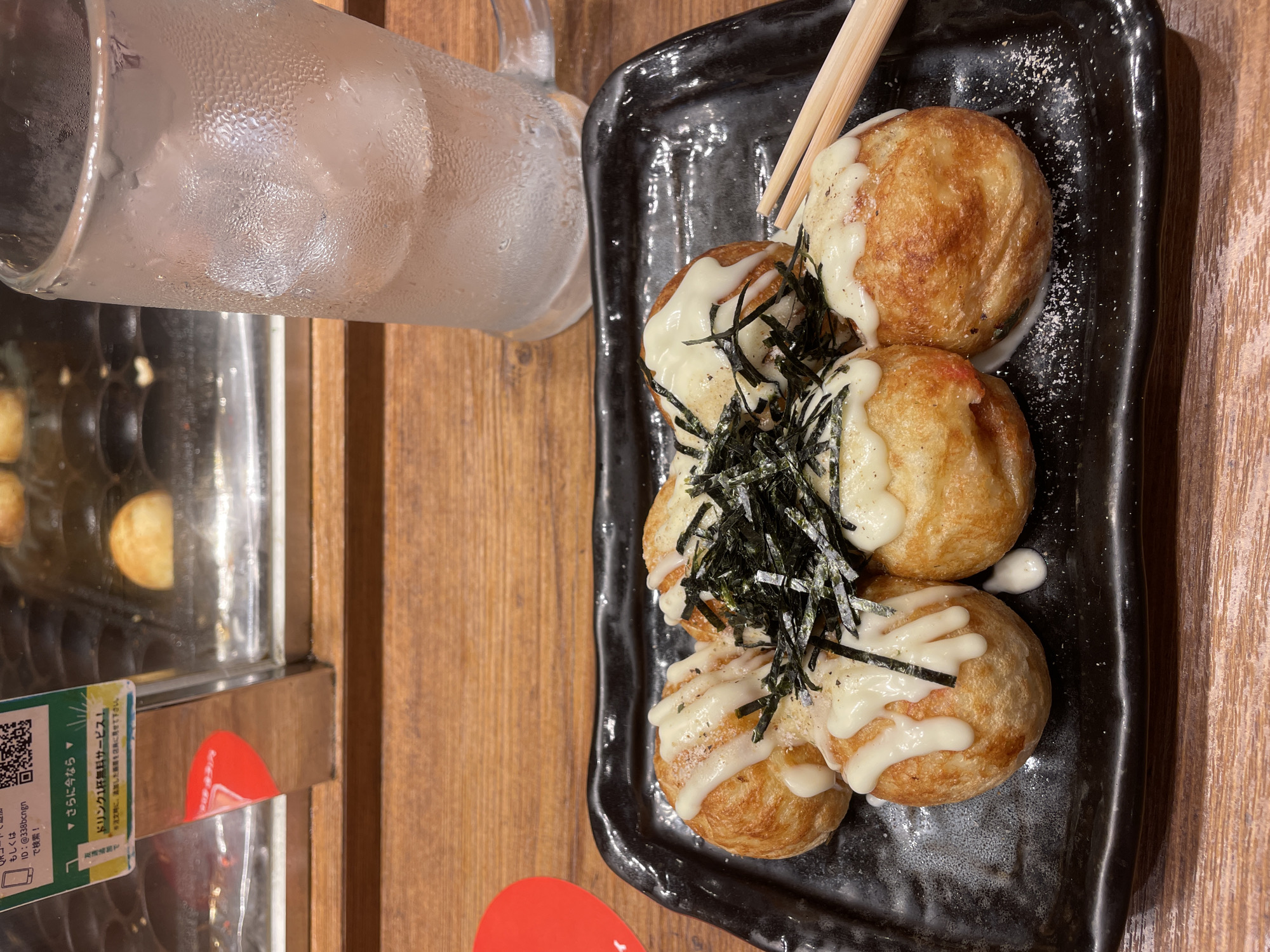 A plate of takoyaki, a Japanese street food, sits on a wooden table, accompanied by a glass of iced water and chopsticks. The takoyaki balls are golden brown and topped with mayonnaise and seaweed. The background shows a food stall with various cooking equipment. The scene is likely from a casual dining establishment in Taito, Tokyo, a district known for its vibrant street food culture.