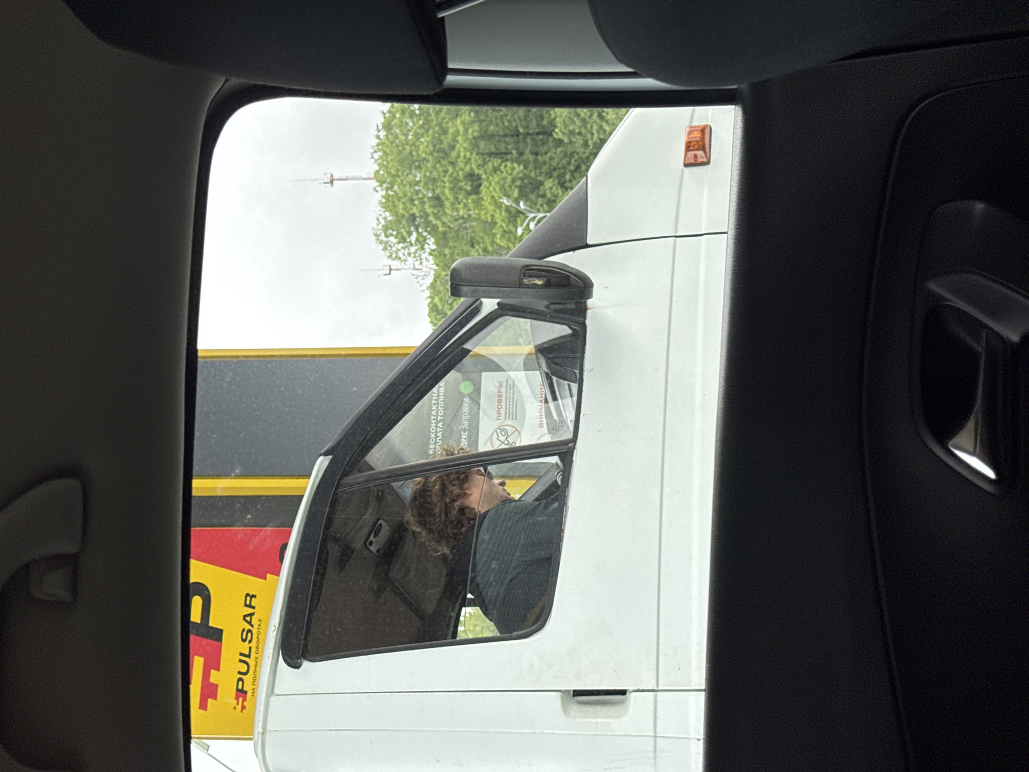 The image shows a view from inside a car, looking out the passenger-side window. A white van is positioned close to the car, partially blocking the view. The driver of the van is visible through the van's side window. He has curly hair and is wearing a dark-colored shirt. Reflected in the van's window are various stickers and documents. A yellow and red advertisement with the word "Pulsar" is partially visible to the left of the van. In the background, beyond the van, there are green trees and light poles. The location is Sochi, Russia.