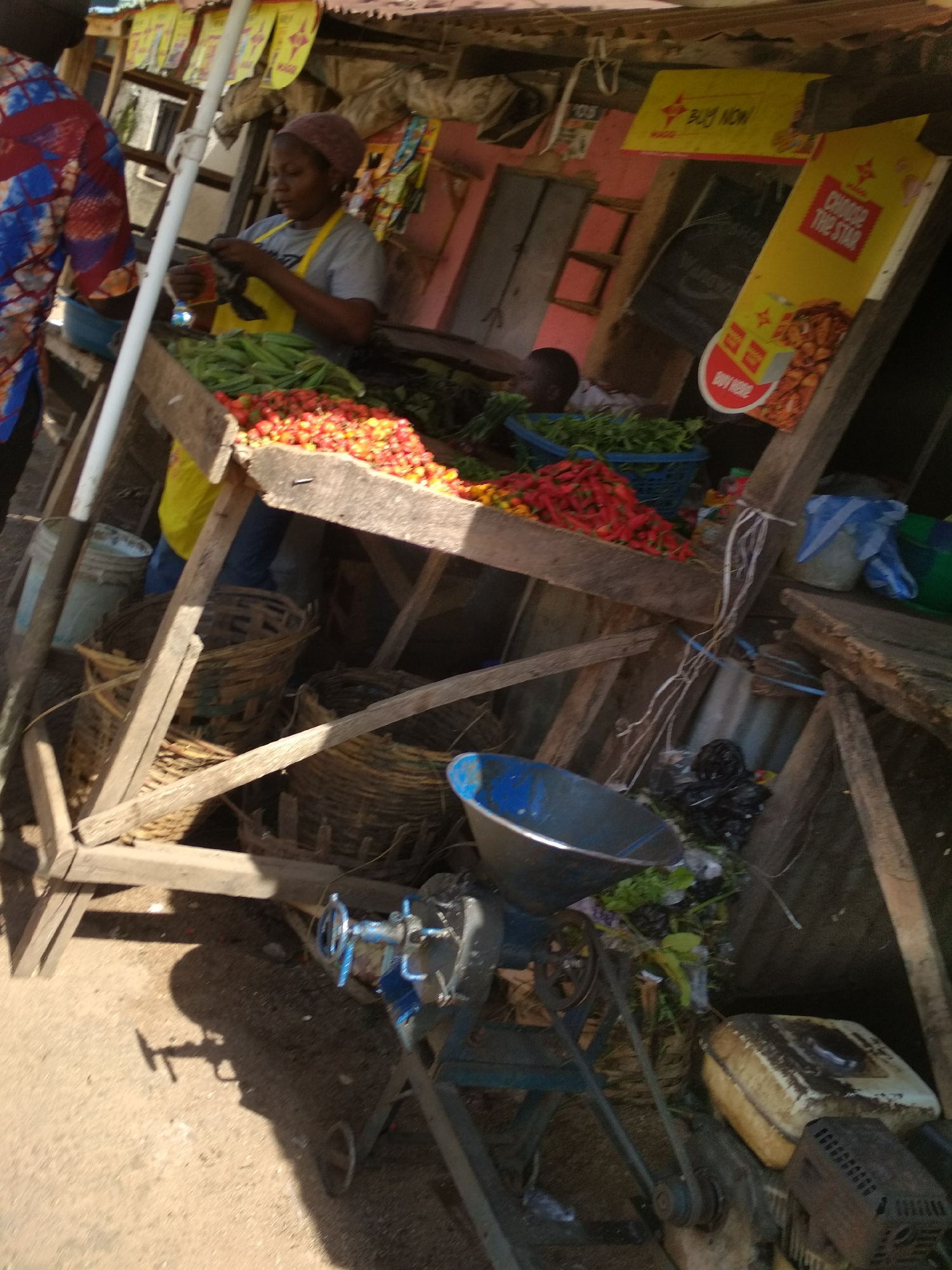 This image captures a vibrant market scene, likely in Jos, Nigeria, focusing on a produce stall under bright daylight. The photograph is slightly tilted to the right.
At the center, a woman wearing a grey t-shirt and a yellow apron, with her hair covered by a patterned scarf, is attending to her stall. She holds a small, dark object in her hands, possibly sorting or preparing items. To her left, the back of another person, dressed in a colorful patterned shirt, is partially visible.
The wooden market stall is rustic, constructed from rough planks and beams. On the stall's surface, piles of fresh produce are displayed prominently: a large quantity of bright red peppers, smaller green peppers, and green okra. Below the counter, several woven baskets are visible, likely used for storage or selling.
Behind the woman, two yellow advertising signs for "MAGGI" are hanging. One smaller sign says "BUY NOW!" while a larger, vertical sign reads "CHOOSE THE STAR" and has a red circle with "BUY HERE" printed on it. In the background, past the stall, a faint figure, possibly a child, can be seen.
On the ground in the foreground, to the right of the stall, sits a blue metal basin and a manually operated grinding machine or mill, with a prominent blue handle. This machine appears to be powered by a small engine, whose fuel tank and casing are visible on the far right. Strong shadows cast on the ground suggest it is mid-day or early afternoon. The scene depicts daily commerce in a bustling, outdoor market environment.