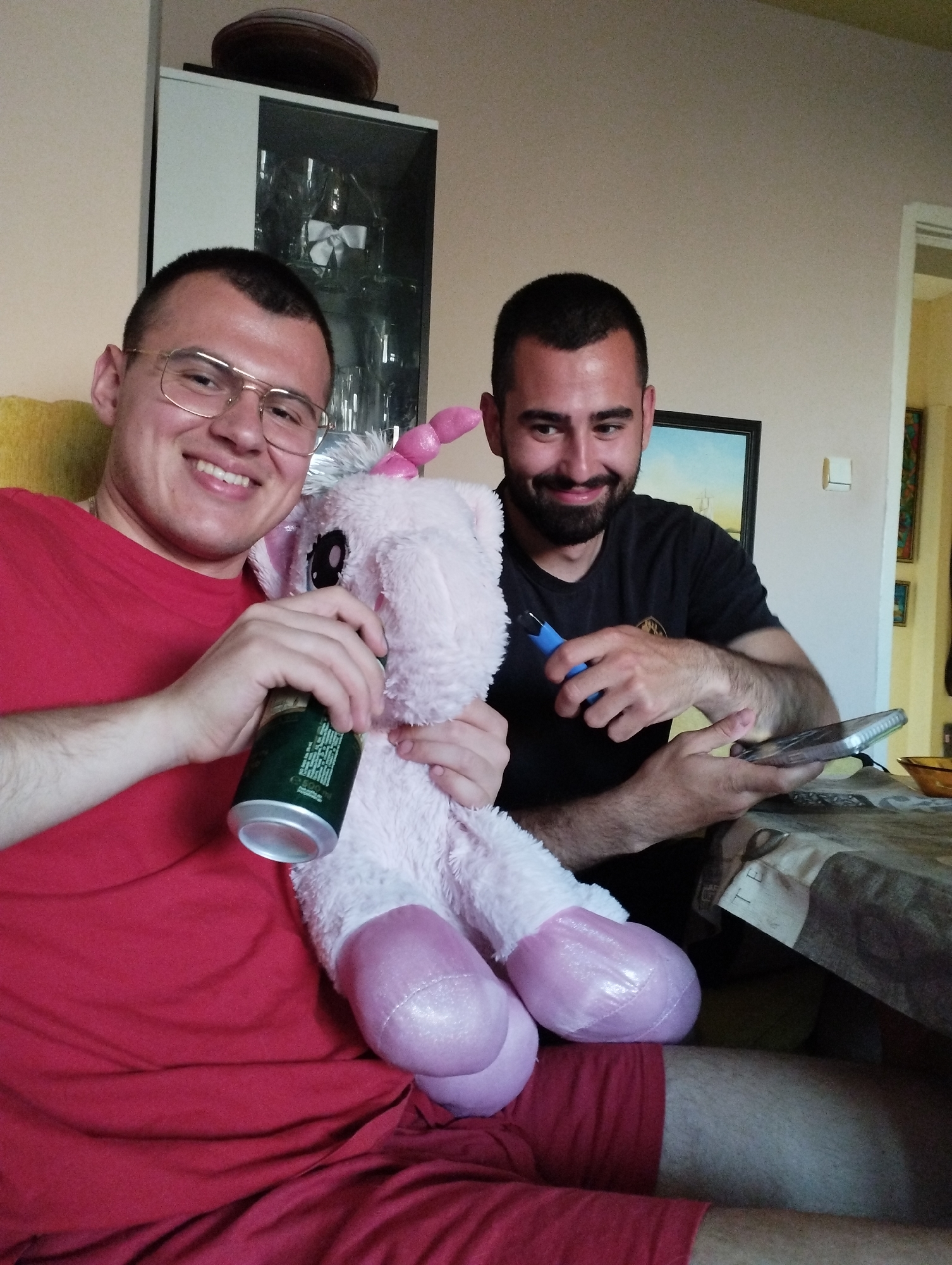 The image shows two men indoors. The man on the left is wearing glasses and a red t-shirt. He is holding a green can in one hand and a pink plush unicorn in the other. The man on the right is wearing a dark t-shirt and has a beard. He is holding a blue tool in one hand and a smartphone in the other. A table is partially visible on the right. In the background, there is a cabinet with glass doors, and a painting on the wall. The location is likely Burgas, Bulgaria.