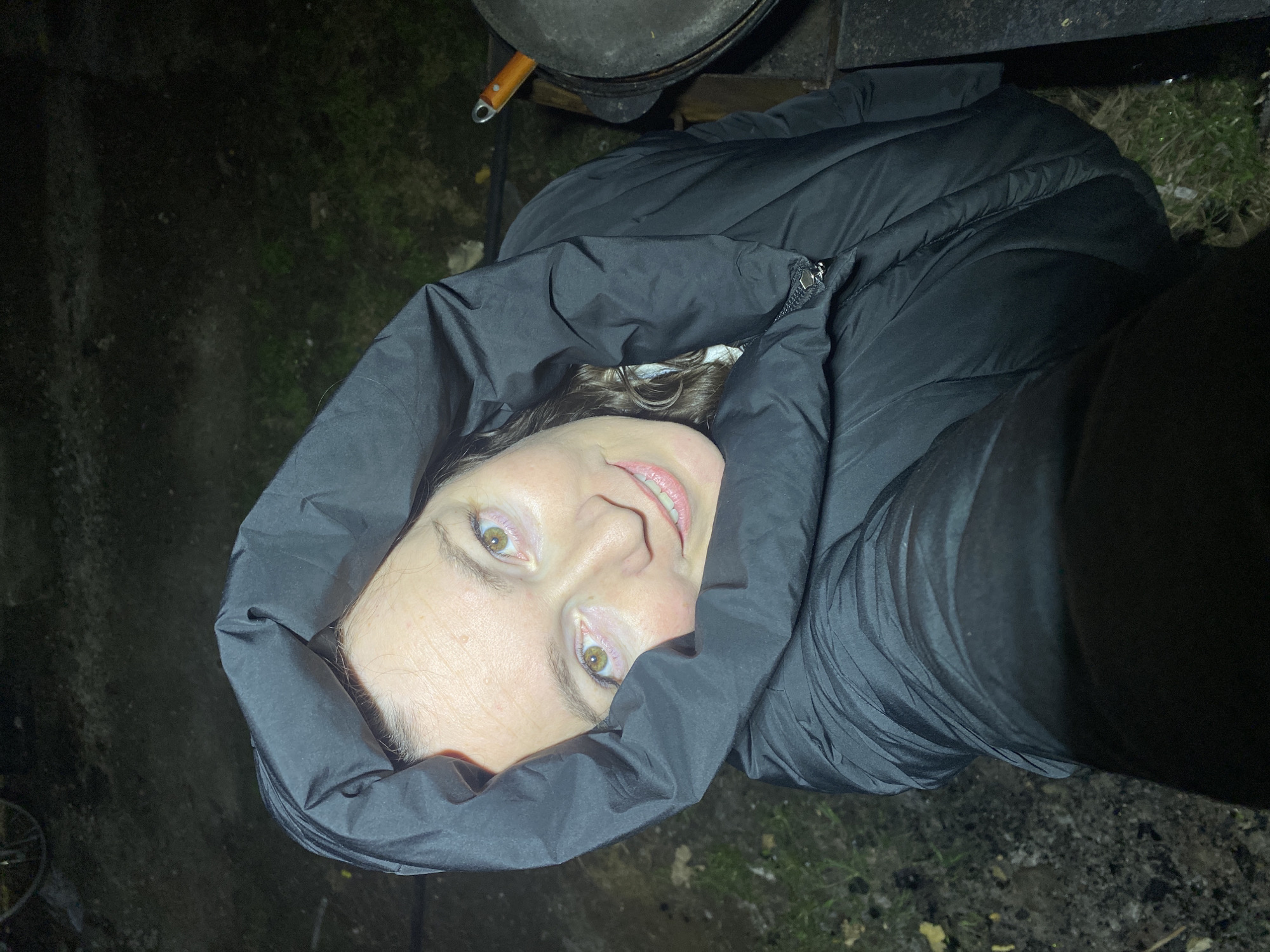The image shows a person, likely female, with light skin, brown hair, and striking green-hazel eyes, captured in a close-up, high-angle shot. Her head is tilted to the side, and she looks directly into the camera with a slight, gentle smile. She is wearing a dark, possibly black, puffer jacket with the hood pulled up, framing her face. A zipper is visible on the front of her jacket.
The setting appears to be outdoors at night or in very low light, with the subject's face illuminated by a direct light source, likely from the camera or a flashlight. The background is dark and indistinct, showing glimpses of uneven ground with sparse grass and dirt, suggesting a rustic or natural environment.
Behind and above the person's head, part of a dark, round object resembling a cast-iron pot or pan with a wooden handle is visible, hinting at cooking outdoors or a fire pit nearby. In the lower left corner, a faint circular object, possibly part of a bicycle wheel, can be discerned. The puffer jacket suggests cool or cold weather. There is no visible text in the image. The location context for this image is Ольховка, Russia.