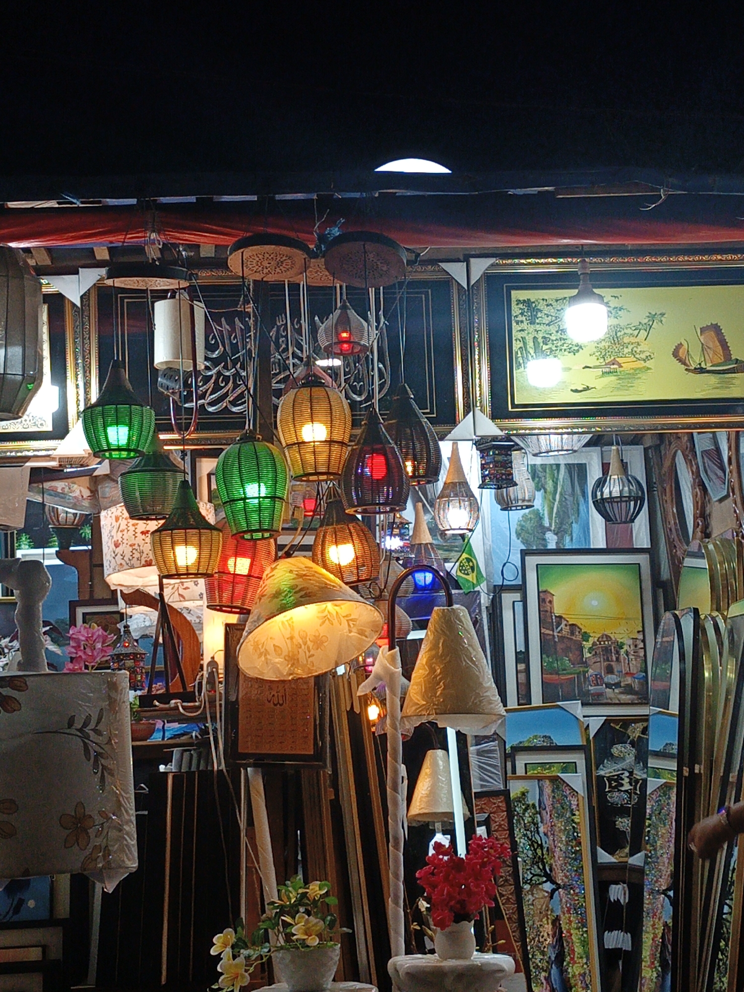 This is a medium shot of an indoor market stall selling a variety of decorative items, primarily hanging lanterns and framed artwork. The lanterns are diverse in color, shape, and material, with many made of woven straw or wicker and illuminated from within. Some are green, yellow, red, or white, and their shapes range from conical to bell-shaped. The artwork displayed consists of framed paintings and prints, with subjects including landscapes, boats, and possibly calligraphy.
The scene is dimly lit, with the primary illumination coming from the hanging lanterns, suggesting it might be evening or a dimly lit indoor space. The overall impression is one of a vibrant, somewhat cluttered market stall, likely an informal setting given the arrangement of goods. The variety of items suggests a place where decorative and handcrafted goods are sold. There is no visible indication of specific weather conditions or time of day beyond the artificial lighting. No people are clearly visible in a way that indicates they are part of an activity, though a hand is visible on the far right of the frame. There is some text visible in the artwork, but it is too blurry to read.