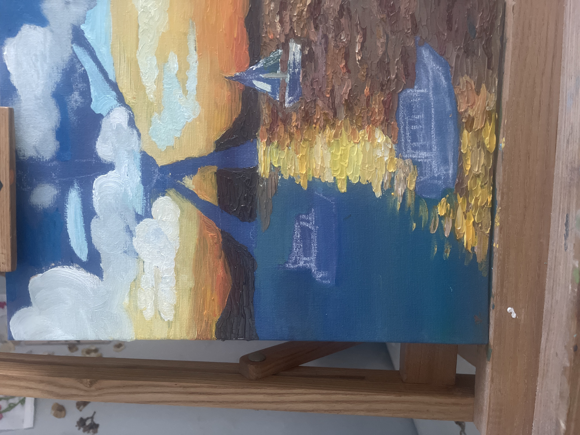 The image shows a close-up of an unfinished oil painting on a canvas placed on a wooden easel. The painting depicts a waterscape with prominent use of textured brushstrokes. In the background, there's a sunrise or sunset with orange and yellow hues. On the left side, there are cloudy formations, painted in white and blue.
In the center, there's a suggestion of land with brown and yellow vegetation. On the water, there are outlines of sailboats or structures, rendered in blue and white. The artwork is still in progress, as indicated by the visible sketch lines in some areas. The setting of the artwork in the city of Maidstone, United Kingdom is not visually discernable from the image.