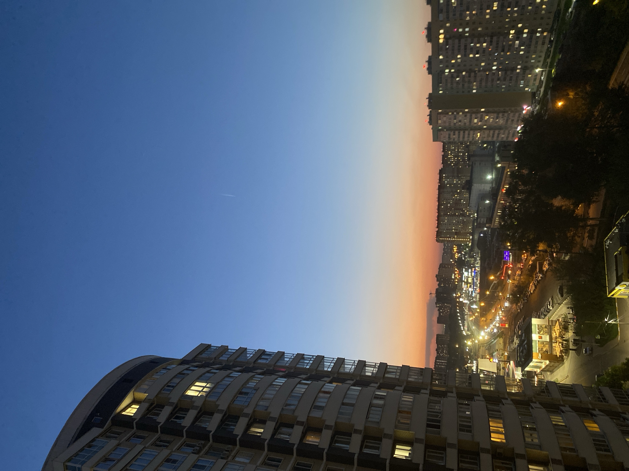 A nighttime view of a cityscape under a gradient sky transitioning from blue to orange. On the bottom left, a curved high-rise building with lit windows dominates the foreground. In the middle right, a city street is illuminated by streetlights and the lights of businesses. In the upper right, other tall buildings with many windows stand in the distance.