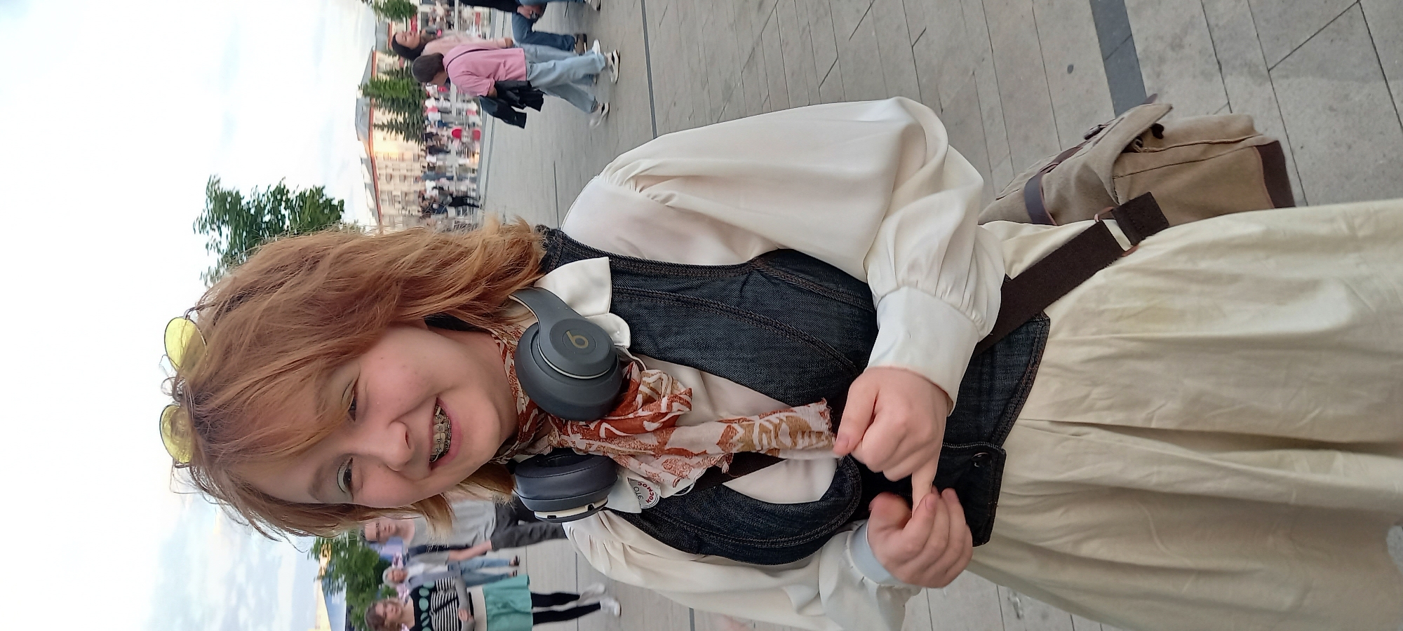 In Izhevsk, Russia, a young woman with reddish-blonde hair, wearing braces and yellow-tinted glasses on her head, is pictured outdoors in a plaza. She has on a white blouse with voluminous sleeves, a dark denim vest, and a patterned scarf. Her hands are clasped in front of her. A grey headset is resting around her neck, and a canvas messenger bag is slung over her right shoulder. The background shows other people walking in the plaza, buildings, and trees, suggesting a public gathering space. The lighting and cloudy sky indicate it is likely late afternoon or early evening.