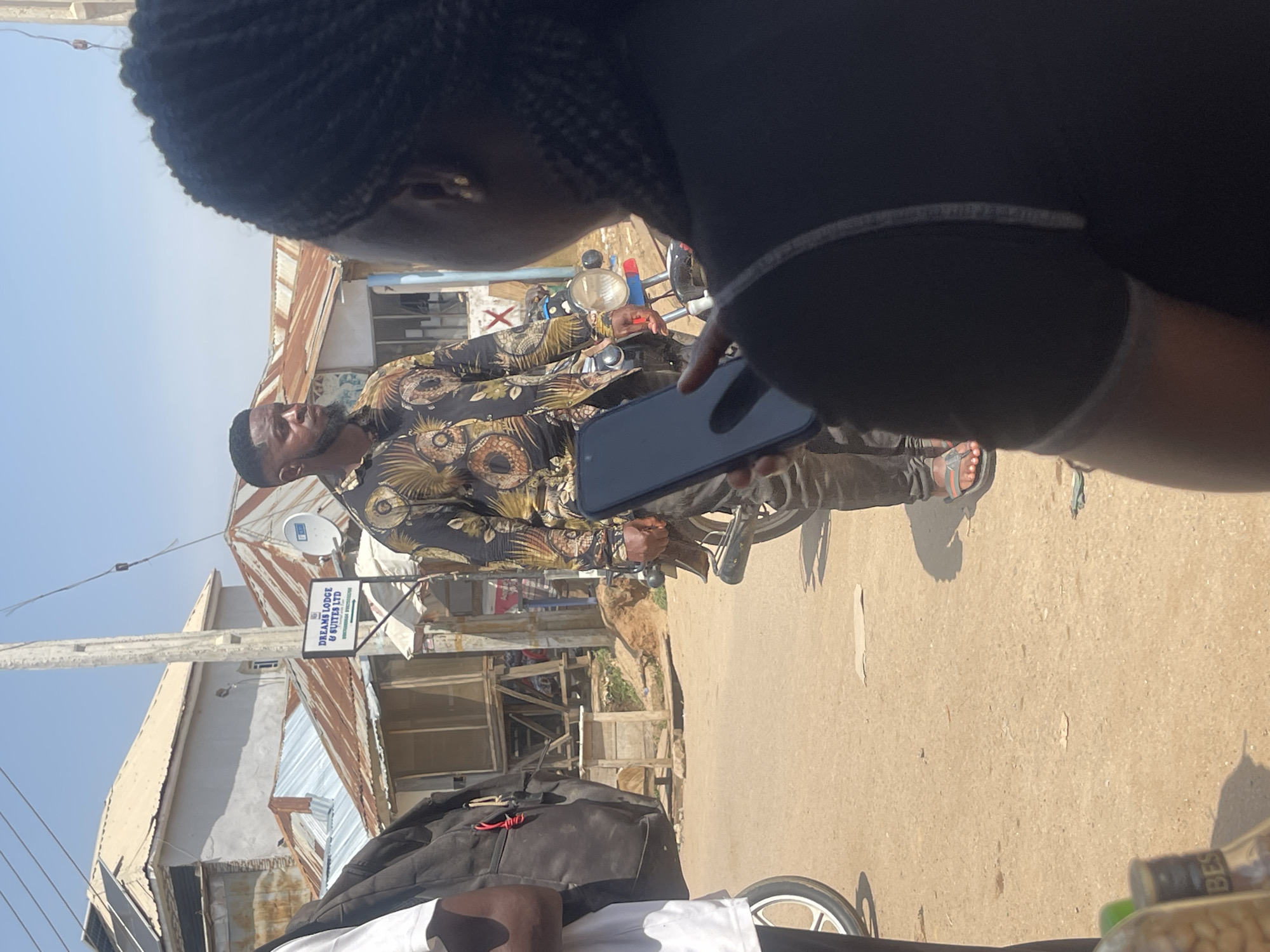 In Abuja, Nigeria, a man with a beard and short hair stands outdoors next to a motorcycle. He is wearing a dark shirt with a gold and brown patterned design and dark pants. He is holding a dark-colored smartphone in his right hand. In the foreground, a person's back is visible, wearing a black top. To the left, a black backpack is seen attached to a person wearing a white t-shirt. The background features buildings with corrugated metal roofs, one of which has a sign that reads "DREAMS LODGE & SUITES LTD." The scene appears to be a street or a marketplace with a dirt ground.