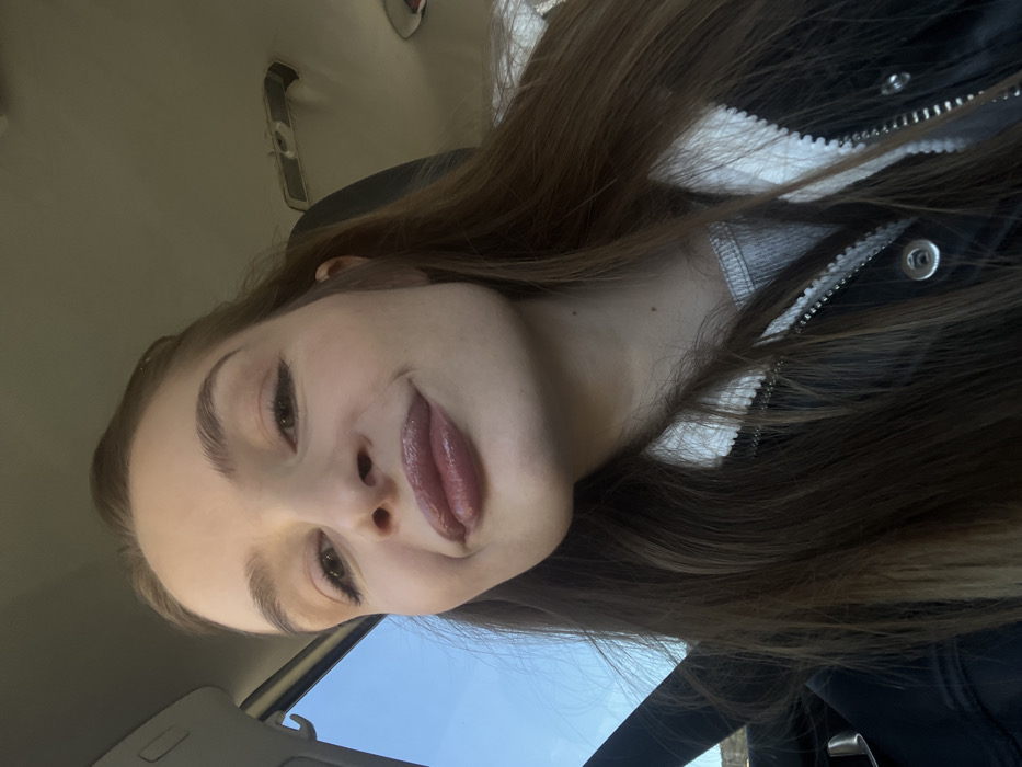 A young woman with long brown hair, glossy lips, and defined eyebrows is captured in an angled, close-up selfie. She wears a dark jacket with a white-lined zipper. The setting appears to be inside a car, evidenced by the vehicle's interior roof and a glimpse of the blue sky visible through the window. The lighting suggests it's daytime.