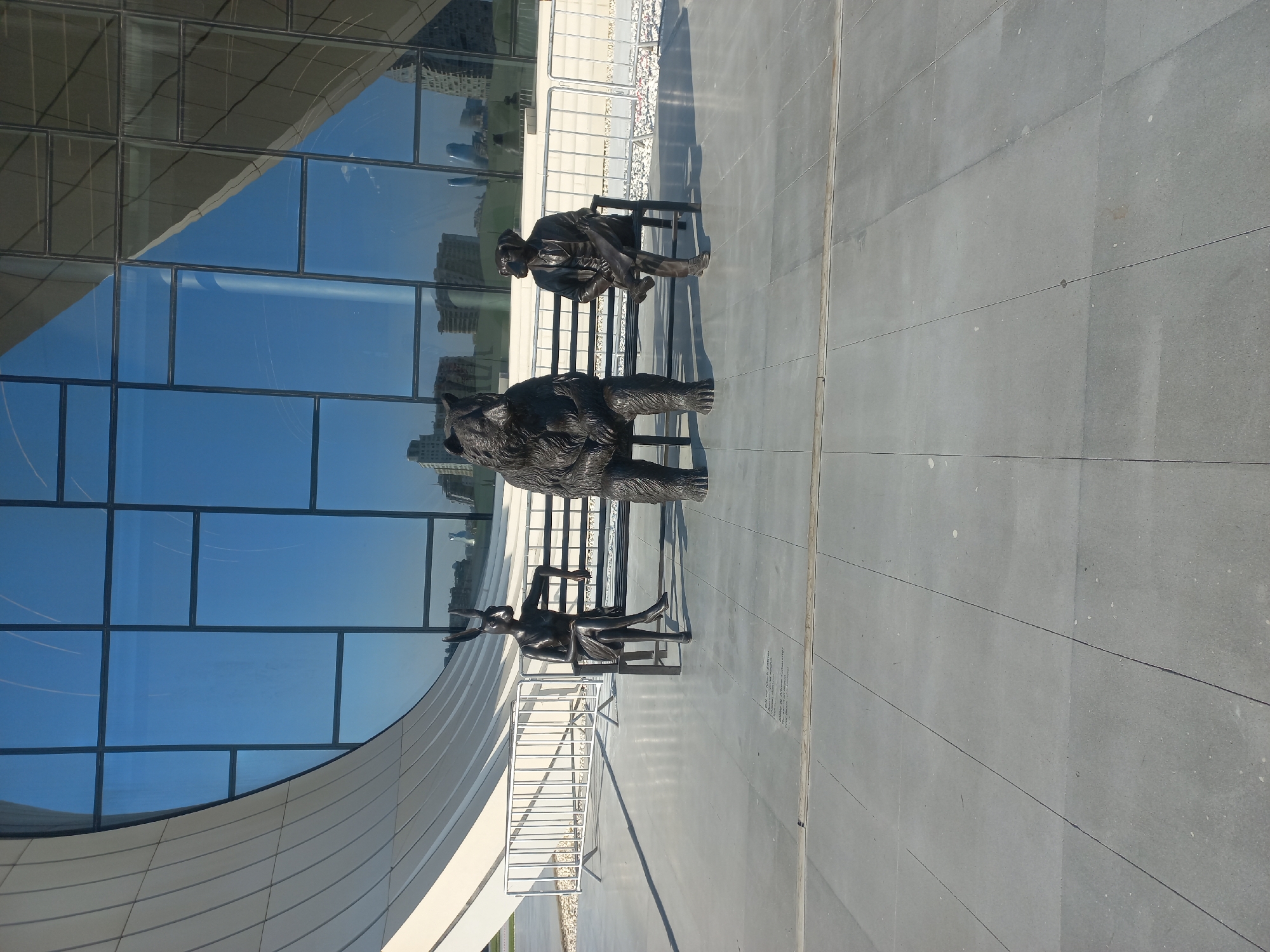 In Baku, Azerbaijan, in front of the Heydar Aliyev Center, three bronze statues are seated on a black bench. From left to right, the first statue depicts a figure with rabbit ears, dressed in a skirt and heels. The middle statue is a large bear, seated upright. The third statue is a dog wearing a suit and tie, holding a newspaper. The statues are positioned on a gray tiled plaza in front of a modern building with a curved, white facade and large, reflective windows. A white metal fence is visible behind the statues, and beyond that, the cityscape of Baku is reflected in the building's glass exterior. The sky is clear and blue.