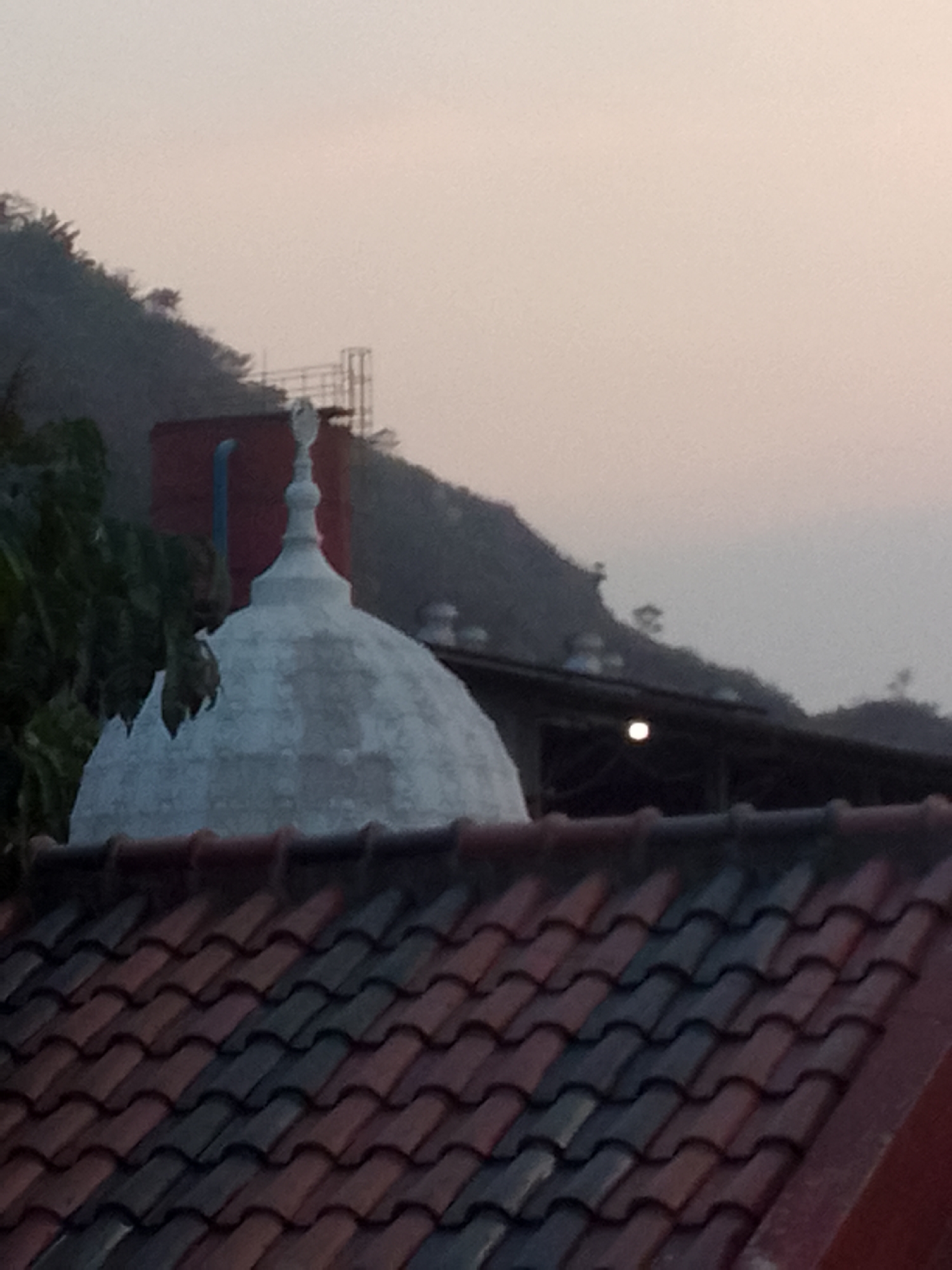 The image shows a rooftop view in the city of Gunungmasigit, Indonesia. In the foreground, a red and grey tiled roof is visible, angled slightly from left to right. Behind the roof, a white dome structure with a pointed finial is prominent. To the left of the dome, part of a red rectangular structure with a blue pipe is visible, partially obscured by dark green foliage. In the background, a densely vegetated hillside rises, shrouded in a light haze. A dark, sloped roof with a visible light source can be seen in the middle ground, positioned between the dome and the hillside. The sky is a pale, washed-out color, suggesting either dawn or dusk. No people or animals are discernible in the frame.