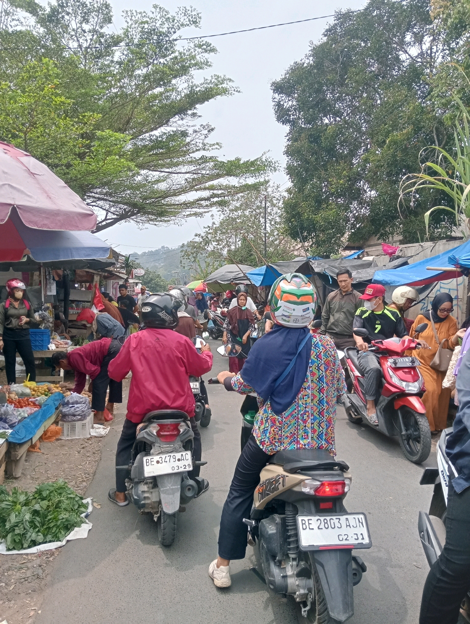 The image captures a bustling scene at an outdoor market in Bandar Lampung, Indonesia. The foreground is dominated by a busy street filled with motorbikes and pedestrians. Several people are seen riding motorbikes, some wearing helmets and face masks. In the background, market stalls are set up along the roadside, displaying various goods, including produce like vegetables and fruits. The stalls are covered with tarps and umbrellas, offering shade from the elements. People are actively engaged in shopping and interacting at the market. The environment suggests a typical day at a local market, with the presence of lush green trees lining the street, indicating a moderately vegetated area. The overall atmosphere is lively and suggests ongoing commerce.