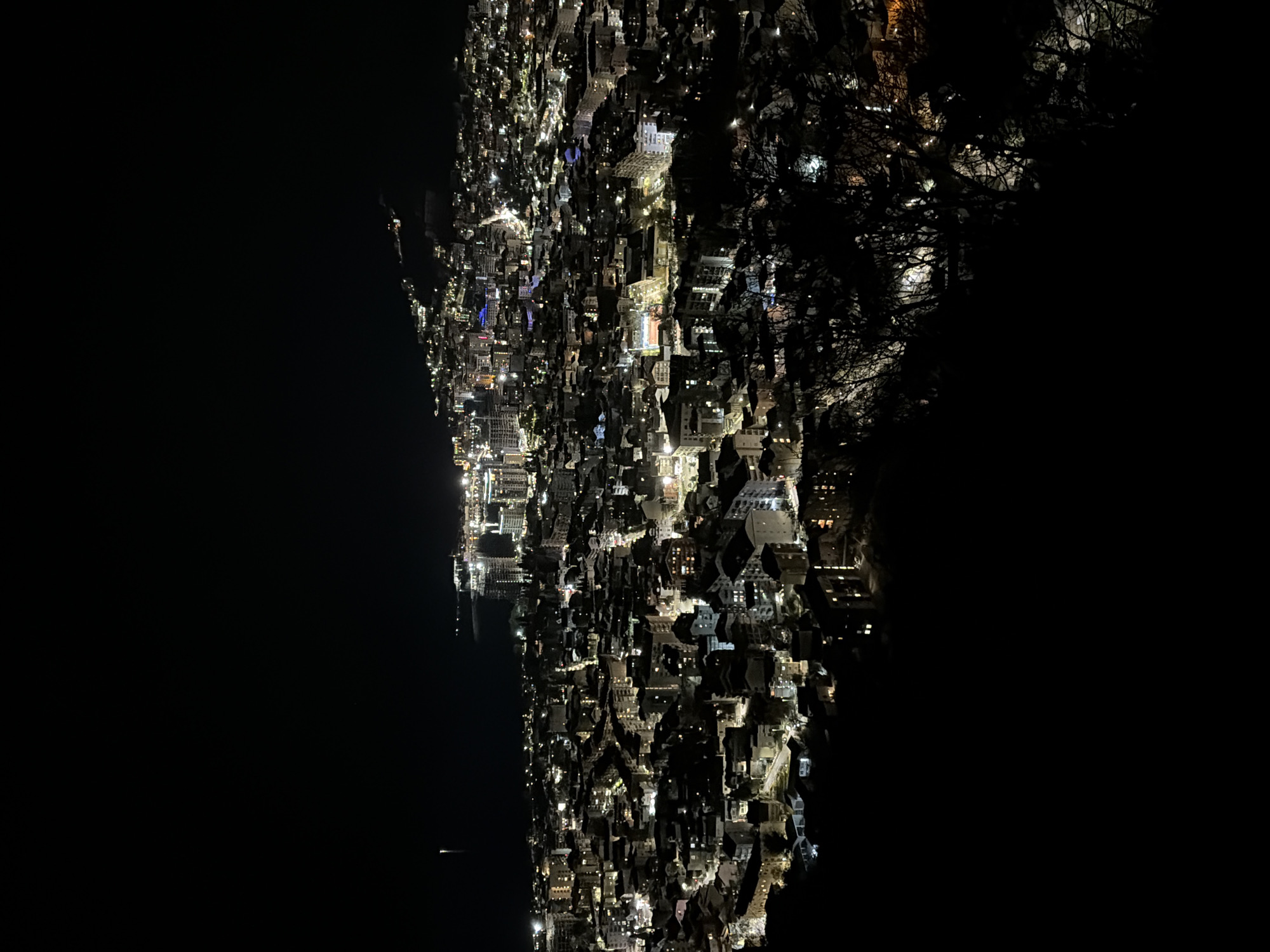 The image depicts a nighttime view of Budva, Montenegro, from an elevated perspective. The scene includes the city’s lit-up buildings, roads, and harbor, juxtaposed against dark surrounding areas. The landscape transitions from a brightly illuminated urban area on the lower half, consisting of closely packed buildings, to a darkened region on the left, indicating the sea and mountains. The upper right area shows a darker landscape, suggesting dense trees or higher ground. The overall impression is of a city at night, tightly nestled between natural elements.