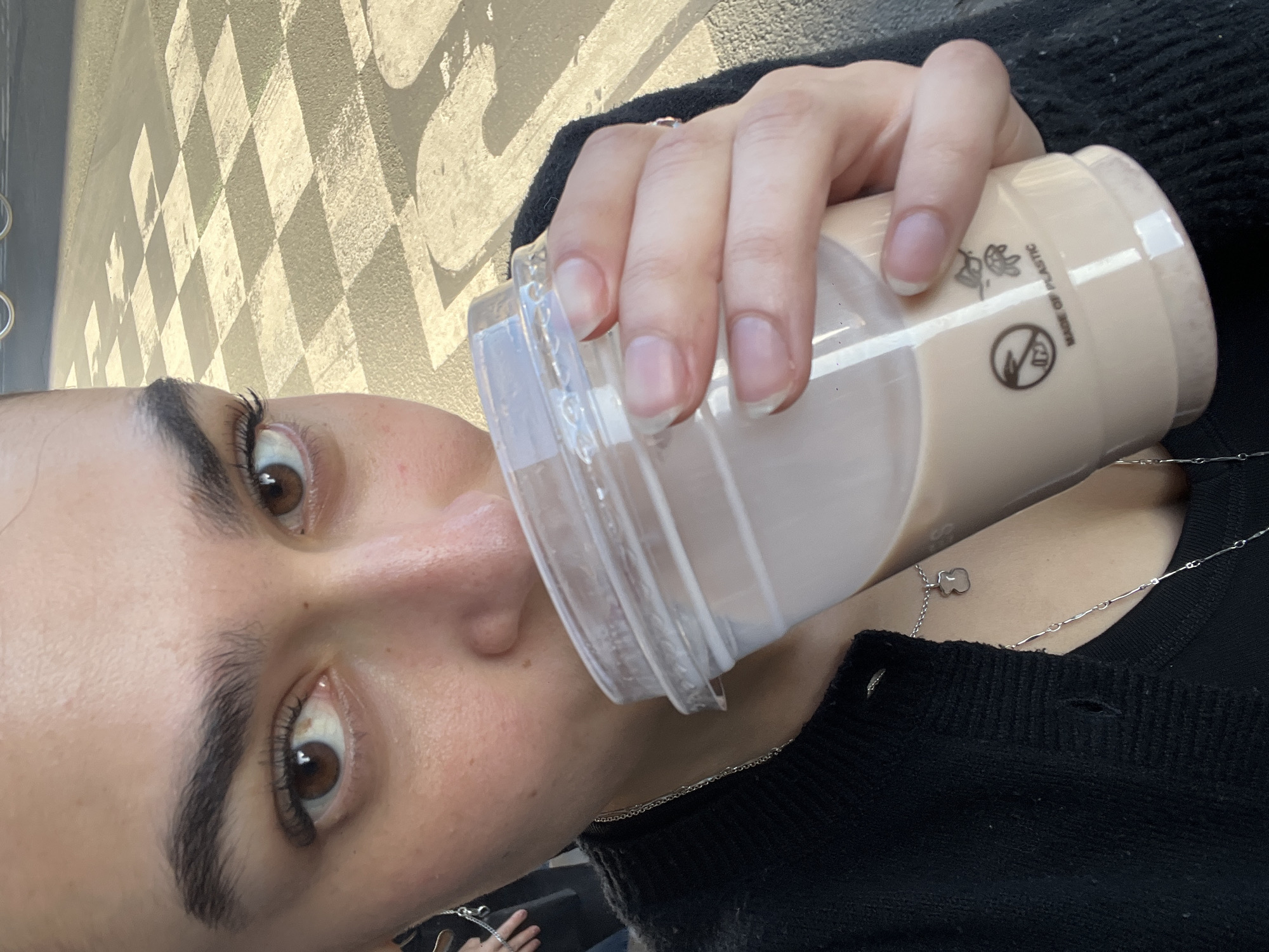 A woman with brown eyes and dark eyebrows is holding and drinking from a clear plastic cup filled with a beige-colored liquid. The cup has a clear lid. The woman is wearing a black sweater, and a silver chain necklace is visible on her neck. In the background, there is a wall or surface with a black-and-white checkered pattern. The location is Paris, France.