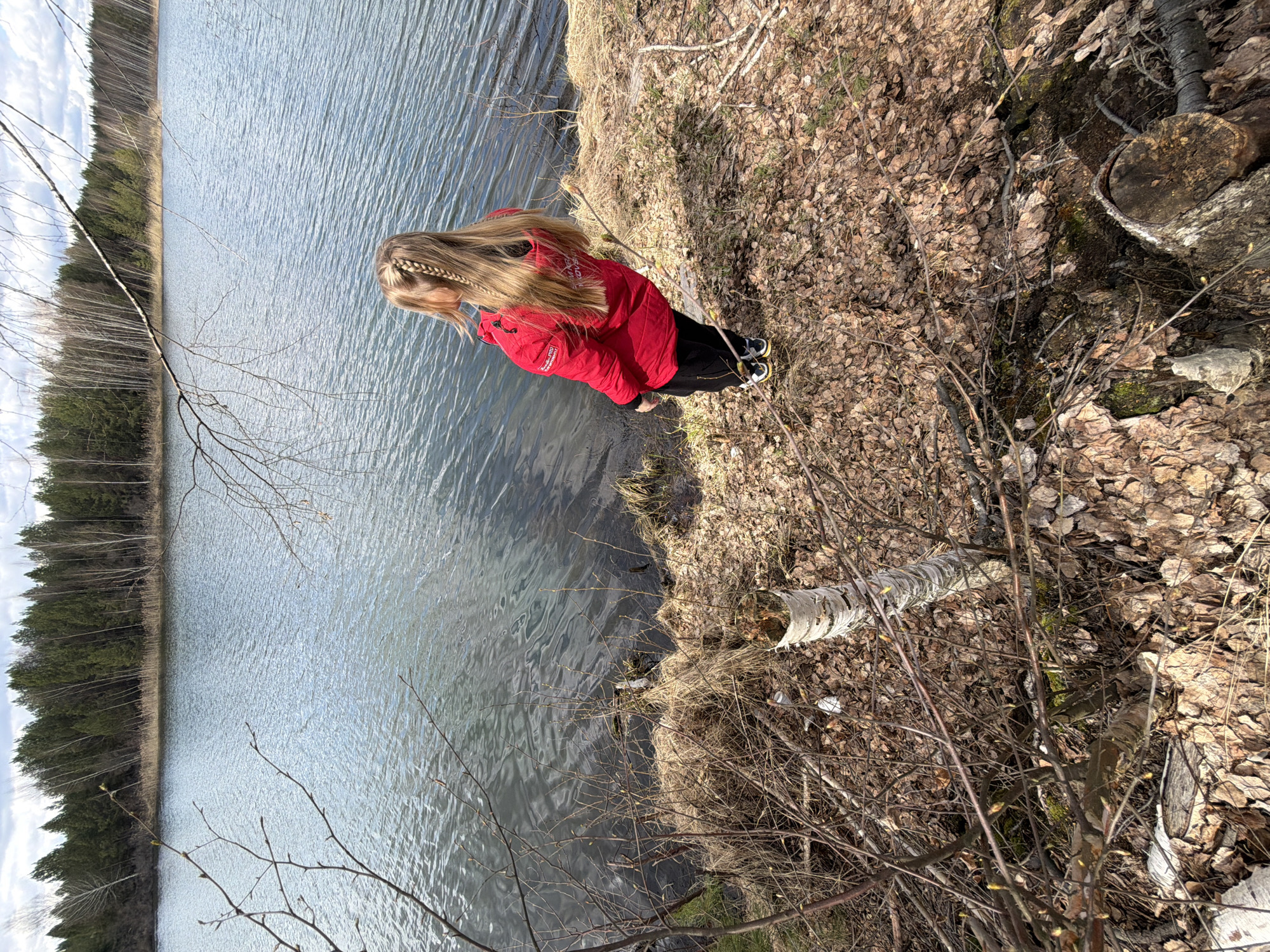 A young girl with blonde hair and a red jacket stands on the edge of a body of water, possibly a lake, in what appears to be a natural setting. The girl is looking down towards the water. The environment includes a forest with bare trees and foliage on the ground. The location is possibly near Novyi, Russia.