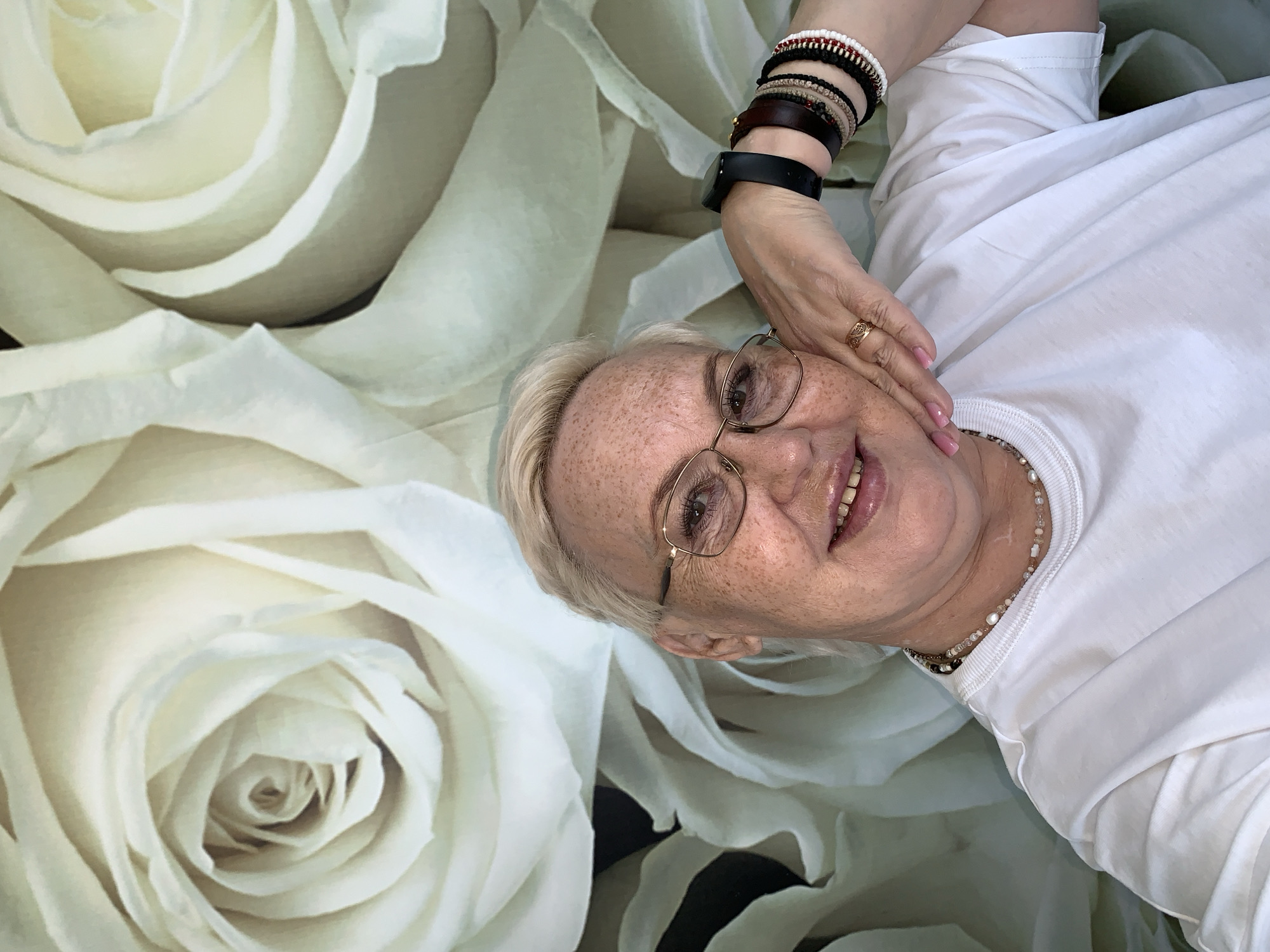 A fair-skinned woman with short blonde hair and freckles poses for a close-up shot on what appears to be a backdrop of large, white roses. She is wearing glasses with a thin gold frame, a white t-shirt, and multiple bracelets on her left wrist, including a smart watch. She also has a beaded necklace. The woman is smiling and her left hand is raised to touch her cheek. There is no visible indication of location in Chaikovsky, Russia.
