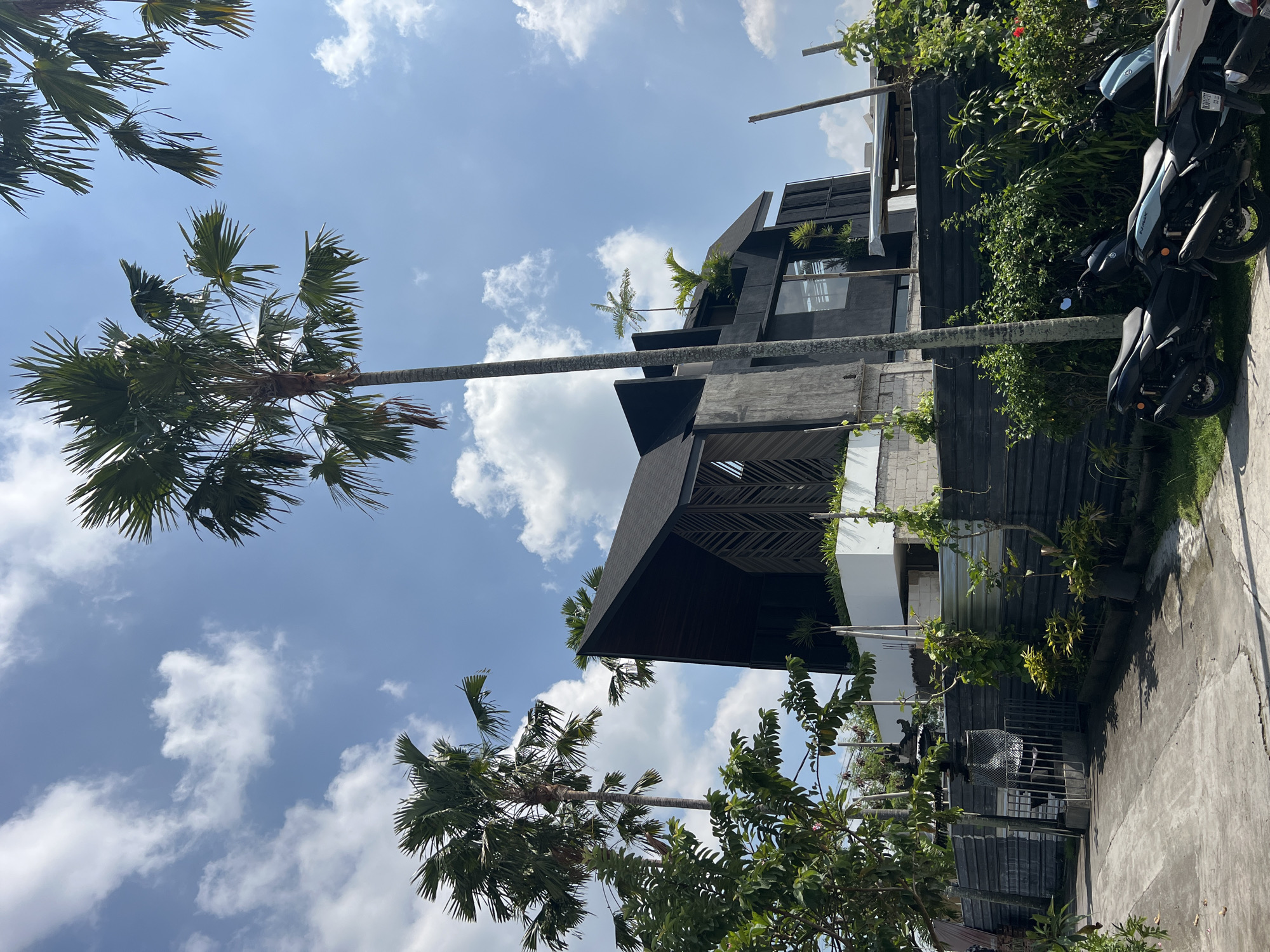 Captured in North Kuta, Indonesia, the image depicts a multi-story modern building with dark facade elements juxtaposed against a vibrant blue sky punctuated by cumulus clouds. Towering palm trees frame the structure, one with a notably slender trunk. The building's architectural features include a partially visible latticed overhang and greenery interspersed throughout its levels. A paved surface leads to the building, where parked scooters suggest recent activity. The photograph, shot from a low angle, emphasizes the building's height and its integration within the tropical landscape.