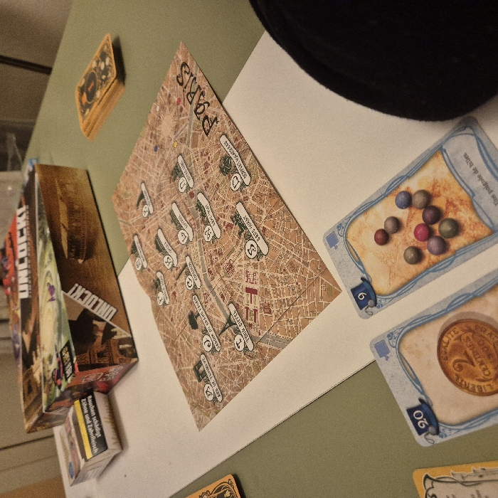 The image displays an Unlock! game setup on a green surface. A brown box of Unlock! is positioned next to a stack of cards. Adjacent to the box is a folded map of Paris, featuring various locations, including the Arc de Triomphe and the Eiffel Tower. Two cards are visible, one depicting a handful of marbles and the number 6, and the other a French coin worth 2 Centimes with the number 20. A black hat sits near the top of the image.