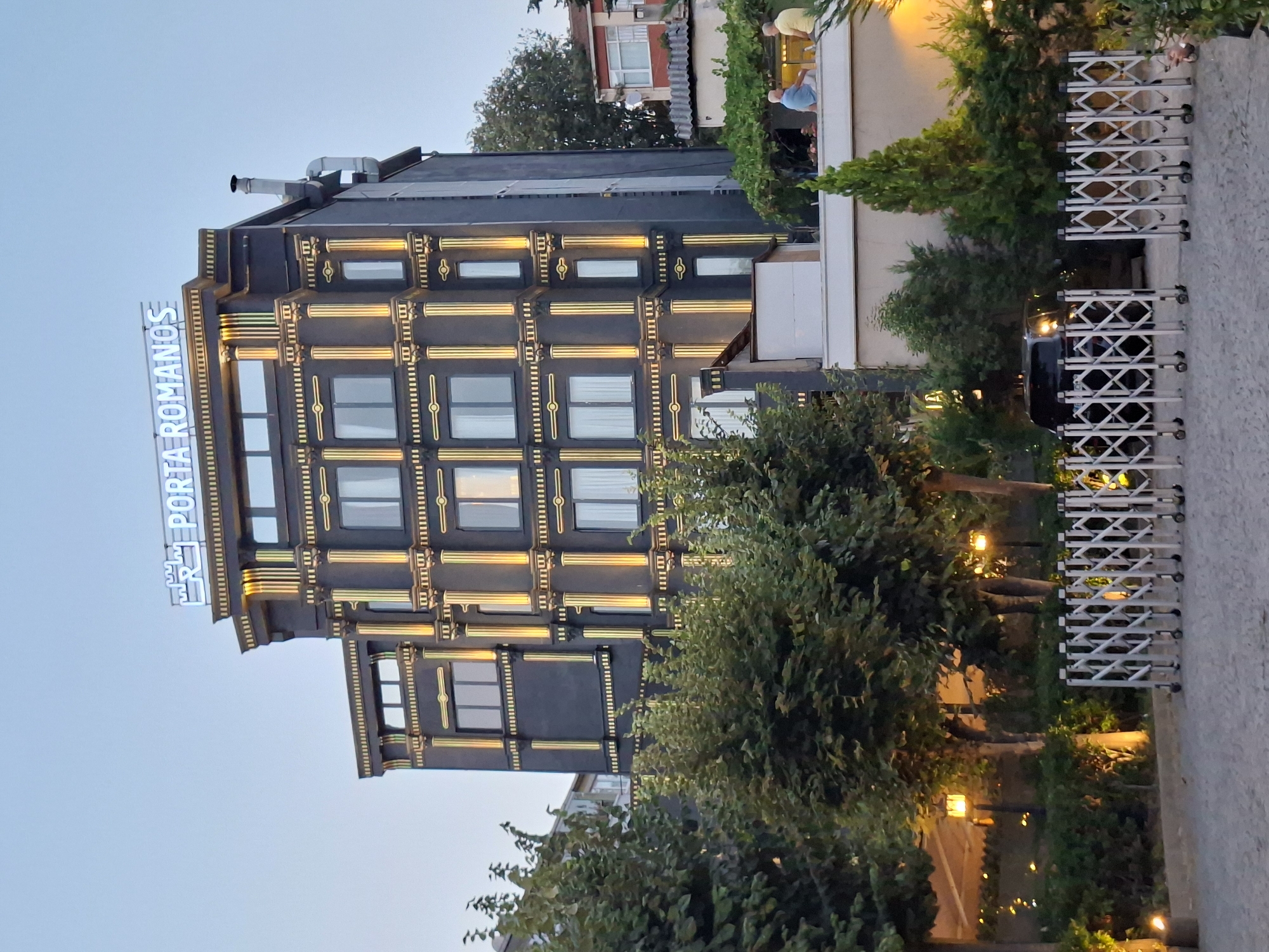 The image depicts the "Porta Romanos" hotel in Istanbul, Turkey, at dusk. The hotel is a dark, multi-story building with ornate gold-colored vertical accents and decorative columns. The windows are illuminated from within, suggesting occupancy. A sign with blue neon lettering reading "PORTA ROMANOS" is visible on the rooftop. In front of the hotel, there is a retractable metal barrier. Beyond the barrier, a dark-colored vehicle is parked. Two men are seated on a rooftop terrace to the right of the hotel, looking outwards. The surrounding area features lush green trees and lampposts, indicating an outdoor setting, possibly a courtyard or street in front of the hotel. The sky is a soft, overcast blue, characteristic of twilight.