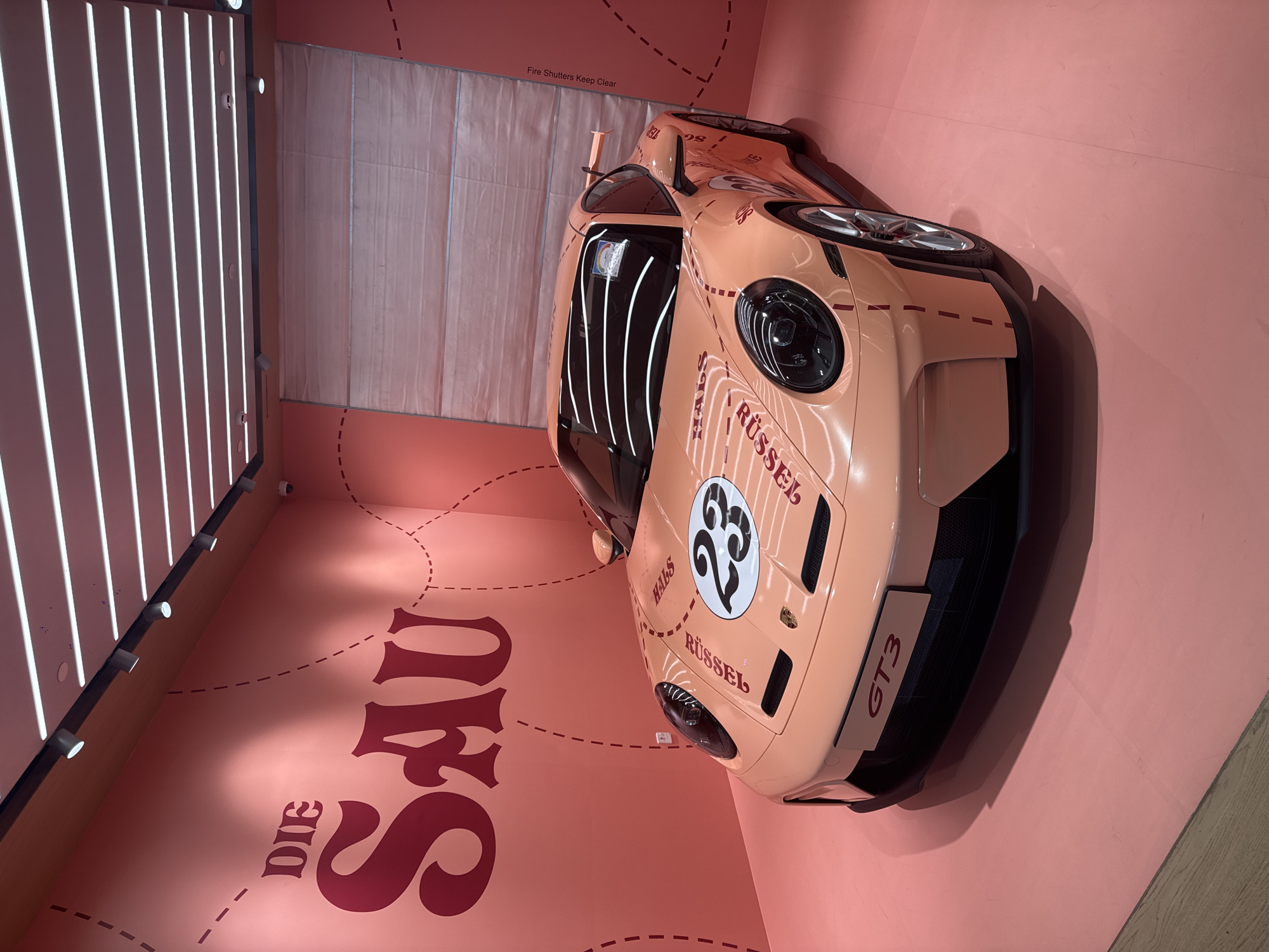 The image depicts a Porsche GT3 in a peach color scheme, placed inside an art installation in Singapore. The car is decorated with a number 23 inside a white circle, and the names "Rüssel" and "Hals" are written on the hood. The phrase "Die Sau" is written in large maroon letters on the wall, and dotted lines create a kind of route or drawing on the wall and floor. A series of vertical light bars are positioned on the wall, and a sign above the back of the car reads "Fire Shutters Keep Clear."