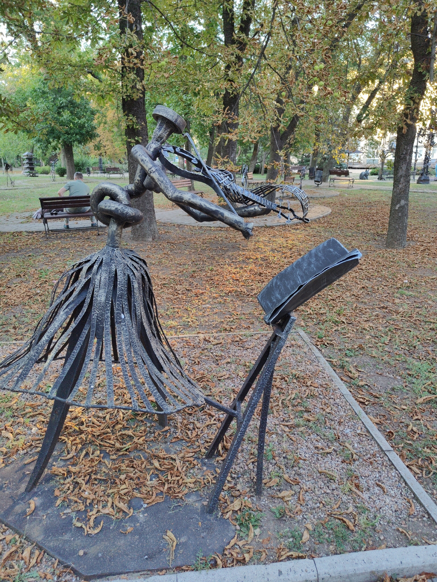 This image shows an outdoor sculpture in a park in Donetsk, Ukraine. The sculpture is made of metal and depicts a figure with a skirt-like base, possibly playing a musical instrument. In the background, there are trees with fallen leaves, benches, and other park elements. A person is sitting on a bench in the distance, facing away from the camera. The ground is covered with dry leaves and gravel.
