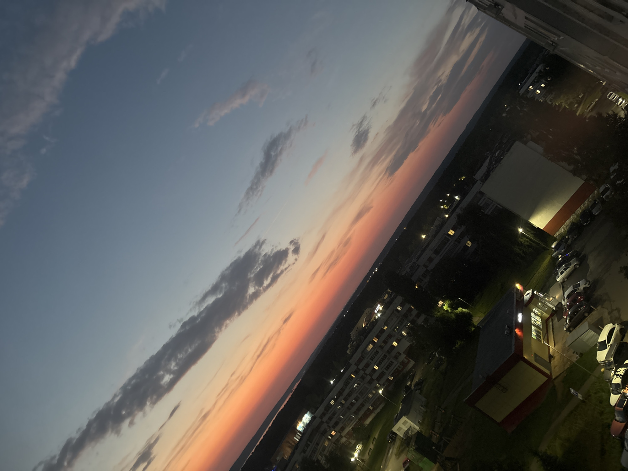 This image captures an aerial view of a residential area in Naberezhnye Chelny, Russia, during sunset. The sky is a gradient of colors, transitioning from a deep orange and pink near the horizon to a pale blue overhead, with scattered clouds. Below, apartment buildings and a commercial building with a red and yellow facade are visible, their windows illuminated. Several cars are parked on the street and in a parking lot. The scene appears to be a typical urban landscape as evening settles.