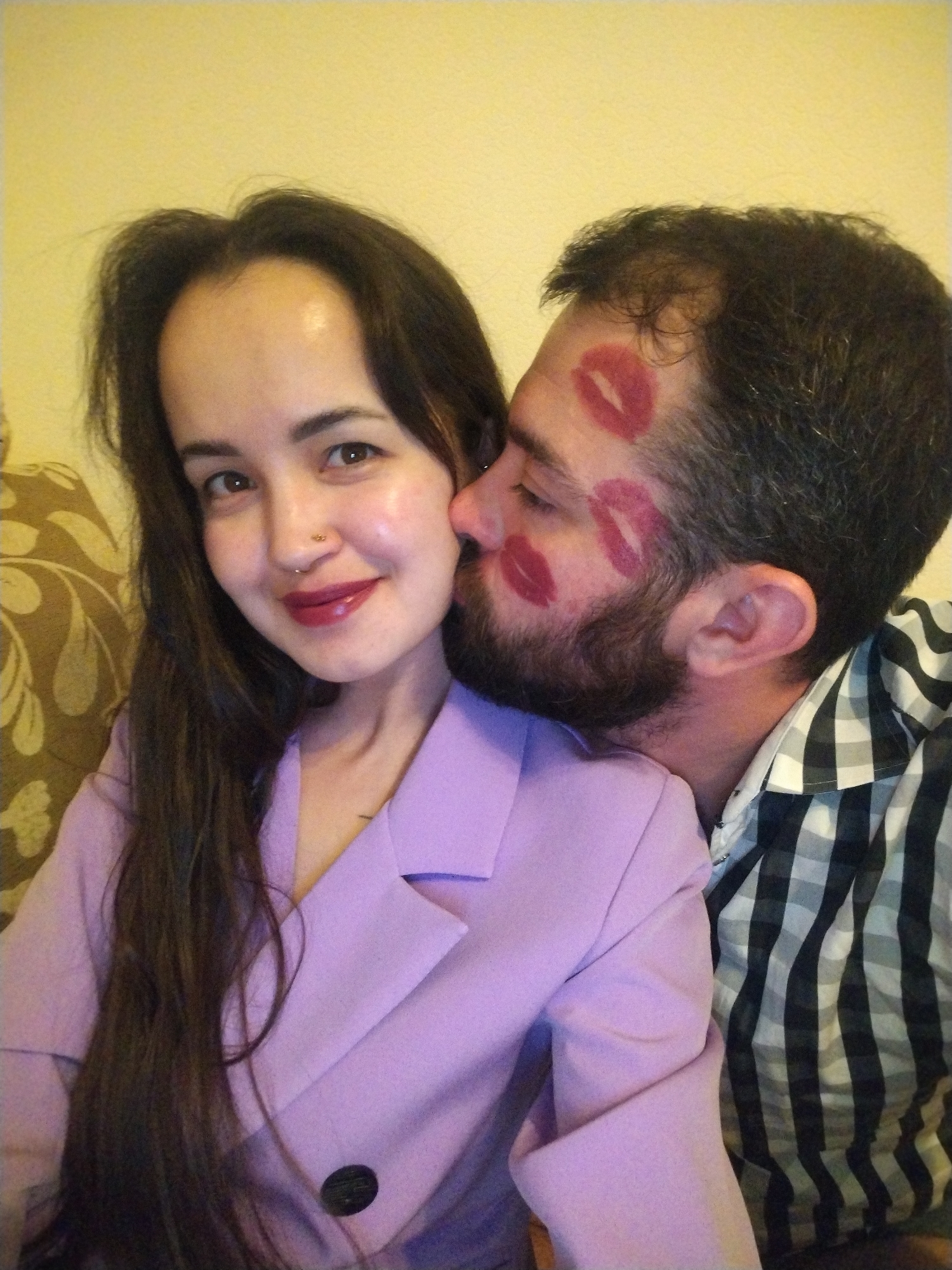 A couple is posing for a photo. A woman with long, dark hair, a nose ring, and dark lipstick is smiling. She is wearing a light purple blazer. A man with a beard has several red lipstick kisses on his face. He is wearing a black and white plaid shirt and has his face pressed against the woman's cheek. The background is a beige wall and a patterned cushion. The image does not contain the name Noviy or any elements associated with Russia.