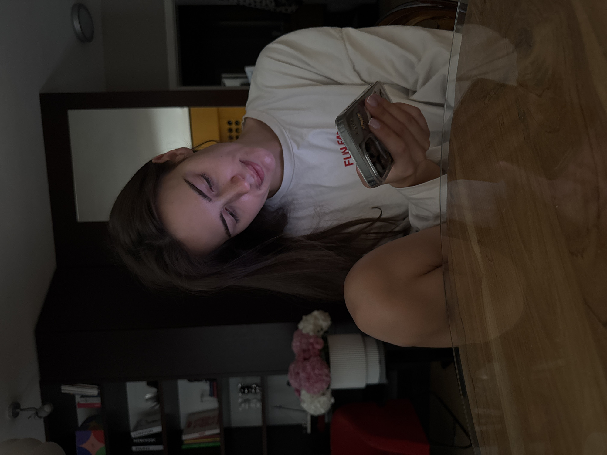 A young woman is indoors, holding a phone with a clear case and the words "FUN FASH" on it. She is wearing a white long-sleeved shirt. She is seated at a clear table that is on a wooden surface. A flower arrangement of white and pink blooms is near her. A dark wooden cabinet filled with books is visible. The photo shows a residential setting with neutral colors and ordinary objects. There is no information in the image to determine that the location is in Warsaw, Poland.