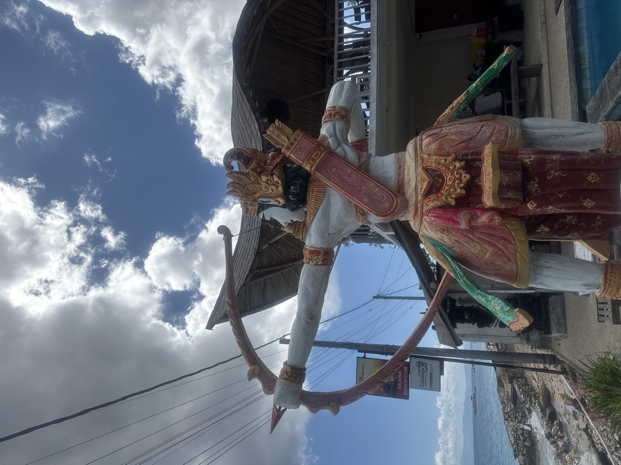 The image shows a statue of a warrior with a bow and arrow, located in Nusa Penida, Indonesia. The statue is painted with white, red, and gold colors and is positioned outdoors, with a building and cloudy sky in the background. There is an Arjuna Beach Club sign visible near the statue. The ocean can be seen in the background. Power lines are visible in the foreground.