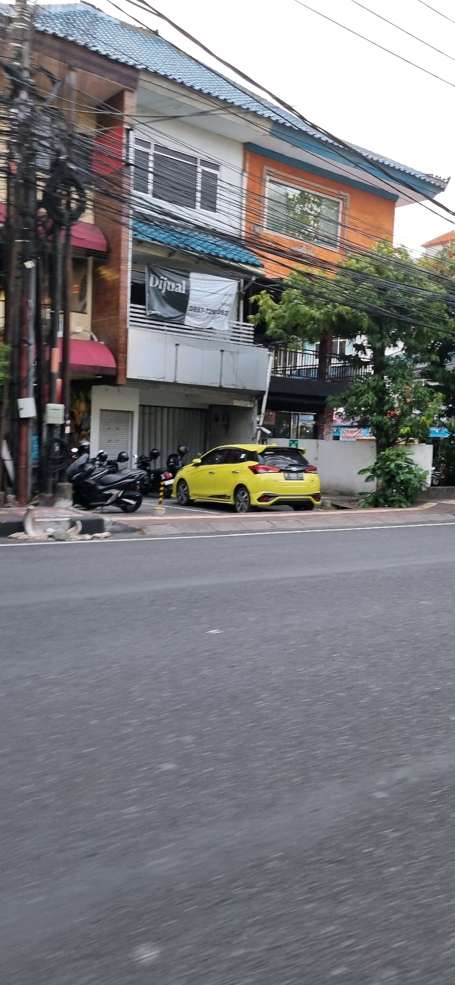 A yellow hatchback is parked on the side of a street in Denpasar, Indonesia. The car is a modern model with a spoiler on the rear. To the left of the car are several black motorcycles parked along a sidewalk. Behind the vehicles is a multi-story building with a blue tiled roof. A banner on the building reads "DIJUAL" which means "FOR SALE" in Indonesian. The building appears to be a commercial or residential property. The street is paved with asphalt and shows signs of wear. Electrical wires are strung across the scene, attached to poles. Trees and greenery are visible behind the building, suggesting a tropical environment. The overall impression is of a typical urban street scene in a Southeast Asian city.