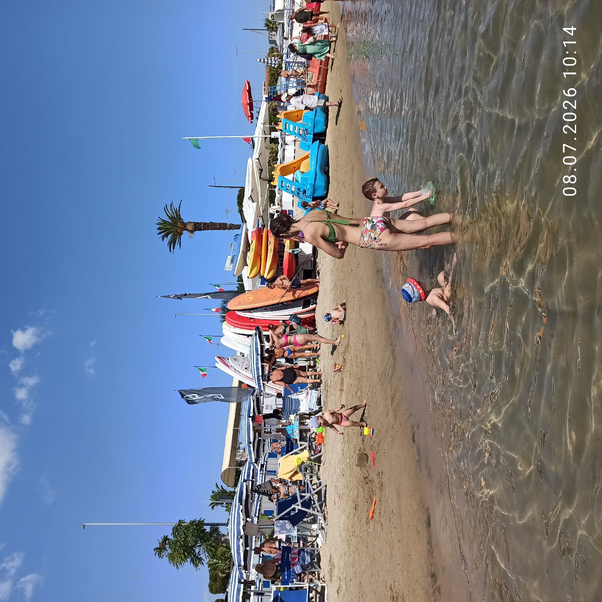The image captures a sunny day at a beach in Trani, Italy, with a group of people enjoying the water and sand. In the foreground, two children are playing near the water's edge. One child, wearing a blue and red hat, is in the shallow water, while another child, a boy, is also in the water nearby. To the right, a woman in a patterned bikini stands with her arms crossed, facing away from the camera. Behind her, the boy is playing with a green object in the water. The beach is lined with rows of blue and white striped umbrellas, providing shade for beachgoers. Several people are seated or standing under the umbrellas. A collection of colorful paddle boats and kayaks are stacked near the shoreline, suggesting recreational activities available at this location. Italian flags are visible on poles amongst the boats and umbrellas. The sand is light brown, and the water is clear with sunlight reflecting off the surface. In the background, buildings and palm trees suggest the coastal town setting of Trani. The date and time stamp "08.07.2026 10:14" is present in the lower right corner of the image.