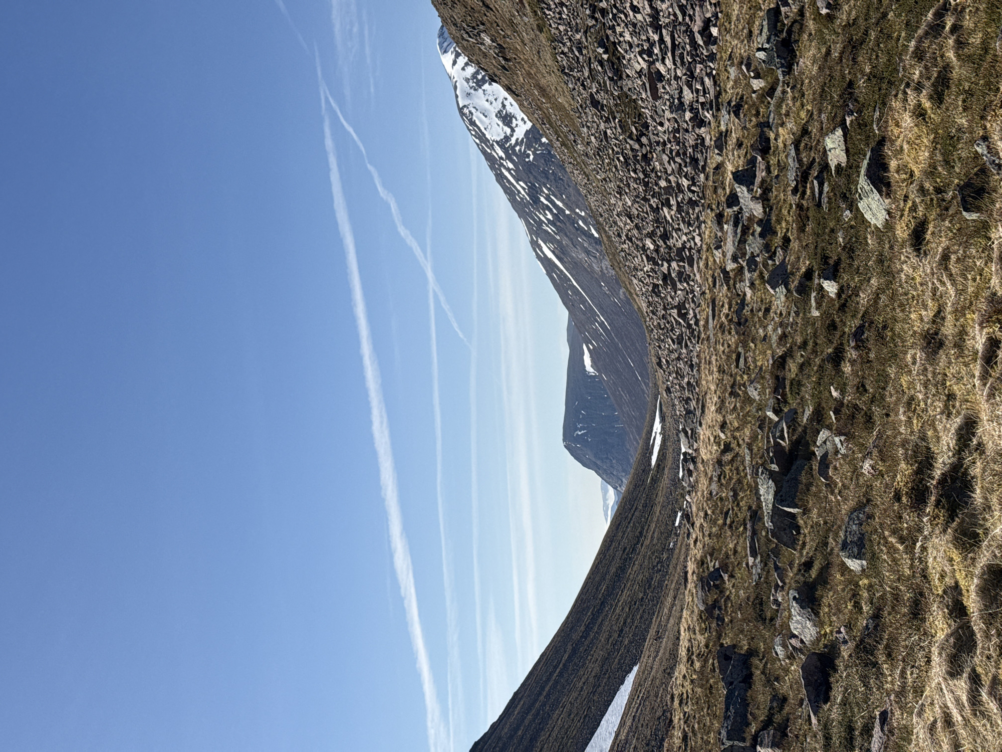 The image depicts a landscape with mountains, a clear blue sky marked by contrails, and rocky, grassy terrain. The mountains in the background are partially covered with snow. The foreground consists of a steep slope covered with a mix of grass and rocks. The picture seems to have been taken in a remote area. There are no signs of human presence or constructions visible. The location is unknown.