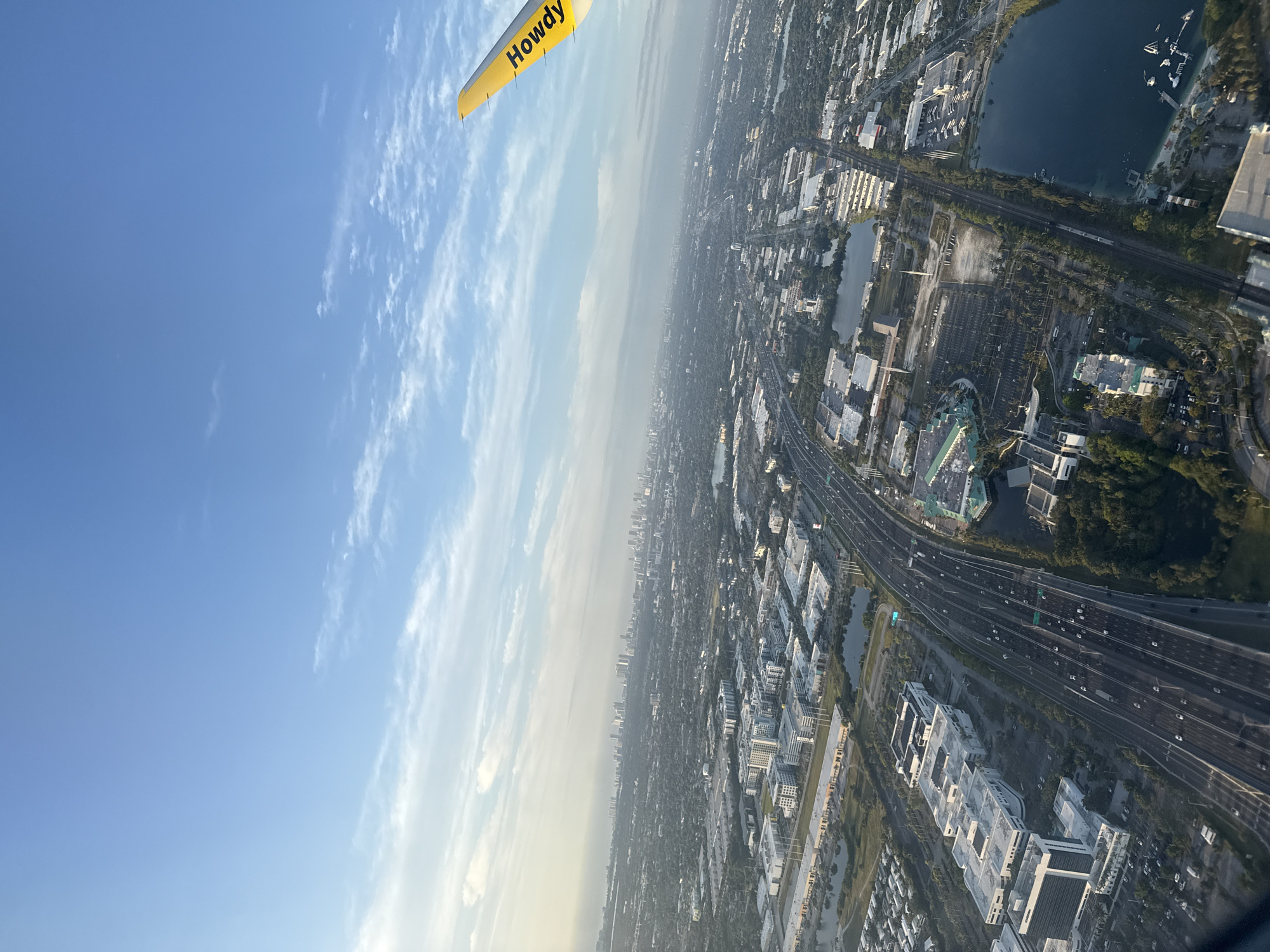 The image presents an aerial view over Dania Beach, Florida, United States of America. A portion of a yellow aircraft wing with the text "Howdy" is visible in the upper left corner. Below the wing is a clear blue sky fading to a hazy horizon. Below the horizon is a cityscape including highways, buildings, lakes, and parking areas. The scene conveys a sense of flight and perspective over a densely populated region.