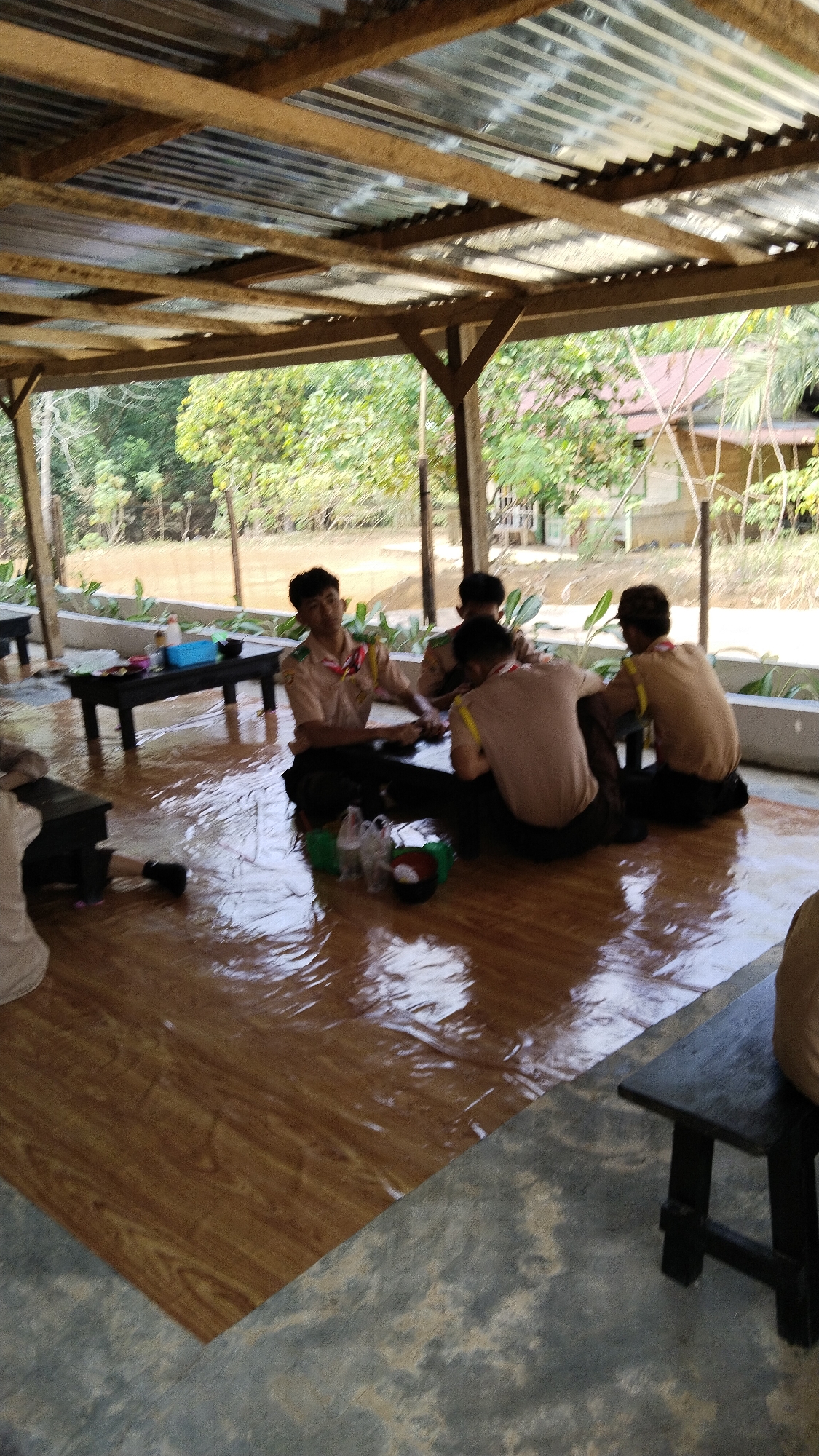 Four individuals, appearing to be young males wearing tan scout uniforms with neckerchiefs, are seated on the floor of an open-air covered structure. The structure has a corrugated metal roof supported by wooden beams and poles. The floor is made of polished wood planks with a visible water sheen, suggesting it is wet. In front of the individuals are two dark wooden tables and a small green bin containing plastic bags and containers. The background shows lush green trees and vegetation, a body of water that appears to be a river, and a building with a red roof. The overall setting suggests an outdoor gathering or activity in a rural or natural environment within Margatunggal, Indonesia.