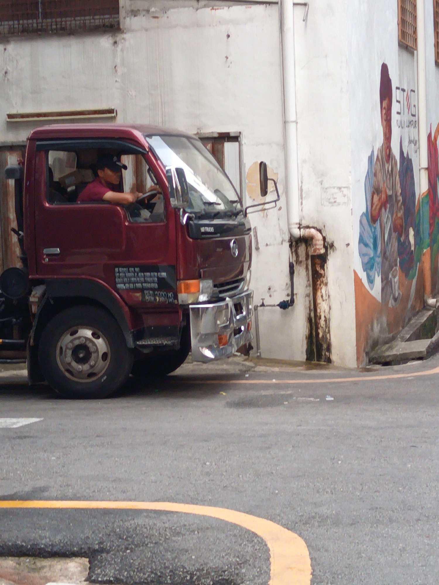 A maroon truck is parked on a street in Kuala Lumpur, Malaysia. The truck has a black steering wheel and a man wearing a maroon shirt and a black baseball cap is visible in the driver's seat. The truck has a large tire on the front left side. The background features a white building with a mural painted on its side. The mural depicts people and the words "STG KUALA LUMPUR". There is a pipe running down the side of the building, and a yellow line is painted on the road in front of the truck. The overall scene appears to be an urban environment.