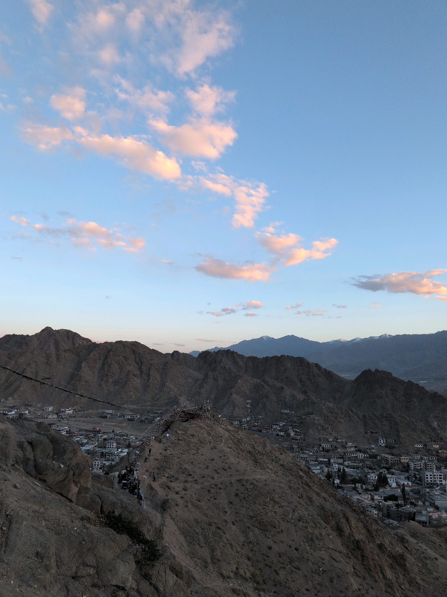 The image shows a panoramic view of Leh, India, from a high vantage point overlooking the city and the surrounding mountains. The sky is a pale blue, with scattered clouds tinged with pink and orange, suggesting either sunrise or sunset. In the foreground, a steep, rocky slope descends towards the city. Below the slope, the city of Leh is nestled in a valley, with numerous buildings, mostly white and light-colored, spread out across the landscape. The buildings appear to be a mix of residential and religious structures, some with flat roofs and others with more traditional architecture.
A winding road or pathway is visible, with a line of people walking along it, suggesting a procession or a popular tourist route. Prayer flags are strung across the path and on the slopes, adding a cultural and spiritual element to the scene. The mountains surrounding Leh are rugged and barren, with dark brown and gray slopes. Some of the distant peaks are capped with snow. The overall impression is one of a remote, mountainous city with a strong spiritual presence. The lighting suggests a time of day when the light is soft and diffused, casting long shadows. The image captures the unique landscape and cultural heritage of Leh.