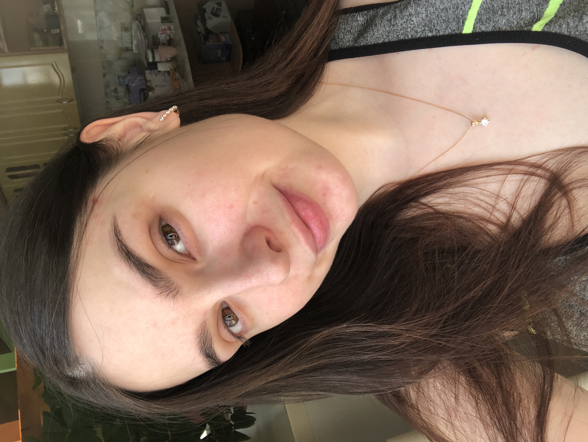 A woman with brown hair and brown eyes is facing the camera with a slight smile. She is wearing a gold necklace with a small diamond pendant and a sparkly earring. She is wearing a gray top with a neon green stripe. The background shows a glimpse of a room with furniture. There is no indication of the location being Noviy, Russia.