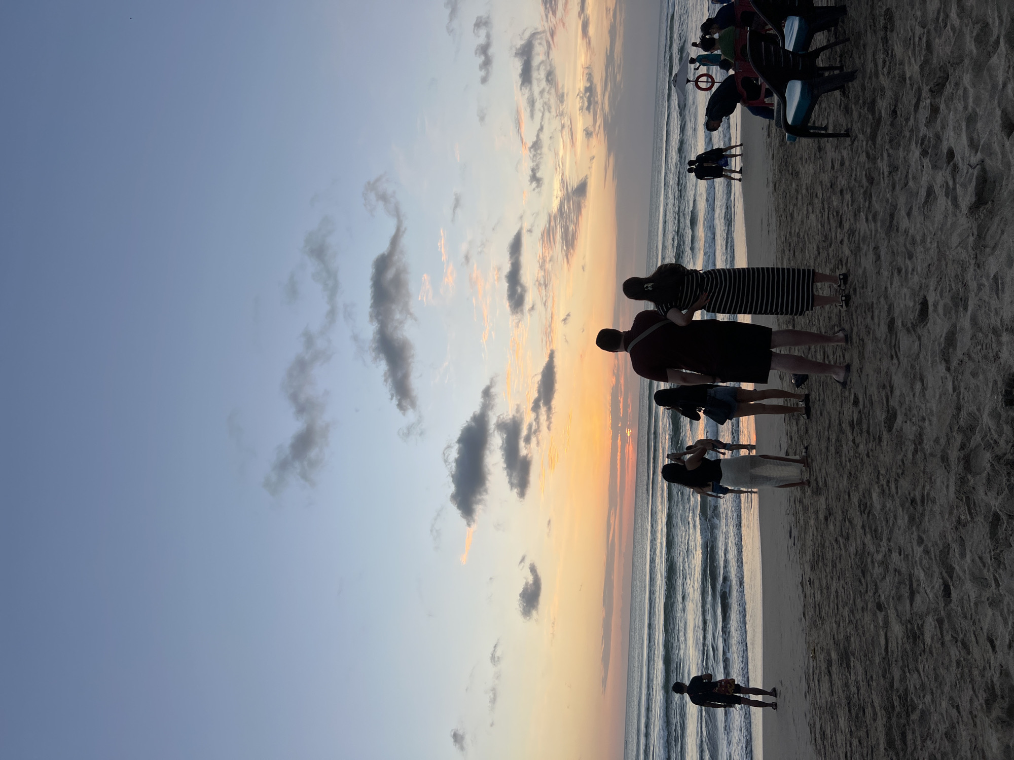 The image depicts a beach scene in Kuta, Indonesia, during sunset. Several people are visible on the sandy shore, some standing and observing the ocean, while others are in the water. In the foreground, a group of individuals are seen from behind, facing the sea. The sky displays a gradient of colors, with hues of orange, pink, and blue, indicative of a setting sun. The ocean has moderate waves, and the beach is covered in sand. Scattered chairs and umbrellas are present further down the beach, suggesting a public recreational area. The overall atmosphere is serene and relaxed.
