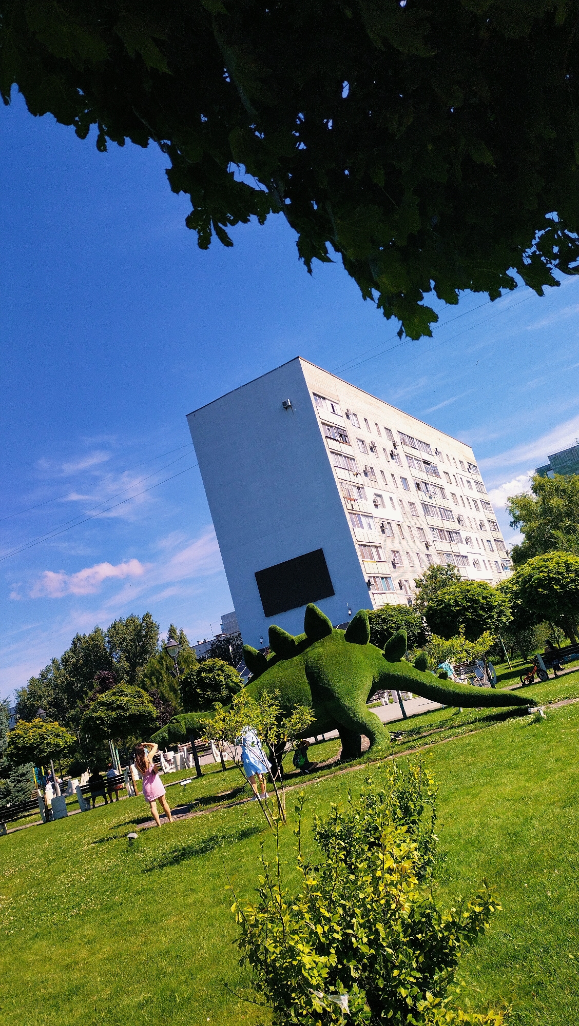 In Horishni Plavni, Ukraine, a large green topiary dinosaur sculpture is prominently displayed on a well-maintained lawn under a clear blue sky. The sculpture, shaped like a stegosaurus with distinct plates along its back, dominates the mid-ground of the image. Behind the dinosaur, a tall, light-colored apartment building with numerous windows and balconies stands at an angle, suggesting a unique architectural design or perspective. The building features a large, dark rectangular panel on its facade.
The park-like setting includes lush green grass dotted with small white flowers, mature trees with dense foliage, and several park benches. Two young women are visible in the mid-ground; one in a pink dress stands with her back to the camera, her head tilted back as if looking at the sky or a nearby tree. The other woman, wearing a light blue dress, is partially obscured by a small tree in the foreground. Other people are scattered in the background, some sitting on benches and others walking. To the right of the dinosaur sculpture, a red electric scooter is parked. Overhead, dark green tree branches with visible leaves frame the top of the image against the expansive blue sky, with faint white clouds present. The overall scene depicts a sunny day in a public park.