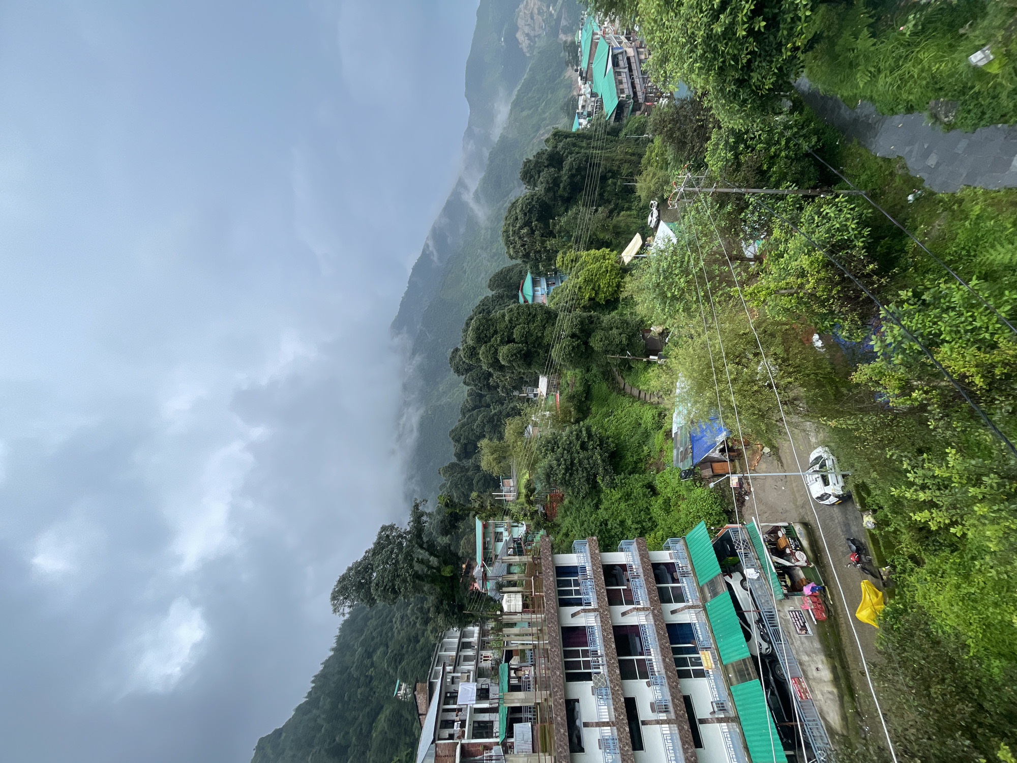 The image depicts a multi-story hotel building with white and brown facades, featuring balconies with blue railings, situated on a lush green hillside in Bhagsu, India. The architecture is typical of hill stations, with several levels of rooms and balconies. In front of the hotel, a road is visible with a white car, a black car, a motorcycle, and a yellow tarp. Some people are present, one holding a pink umbrella, suggesting it might be raining or has recently rained. Green awnings cover the ground floor of the hotel, likely housing shops or restaurants.
Beyond the hotel, the landscape transitions into a dense forest of tall trees, characteristic of the Himalayan foothills. The mountains in the background are shrouded in mist and clouds, creating a dramatic and atmospheric scene. The sky is overcast, with heavy, grey clouds dominating the upper portion of the image. The overall impression is one of a slightly misty, possibly damp, day in a picturesque mountain town. The presence of the hotel and the road indicates a developed area, while the surrounding greenery emphasizes the natural beauty of the location. The style of the buildings, with their distinct green roofs visible further up the hillside, also suggests a regional architectural influence. The image captures a serene yet vibrant atmosphere, hinting at the life and activity within this hillside community.