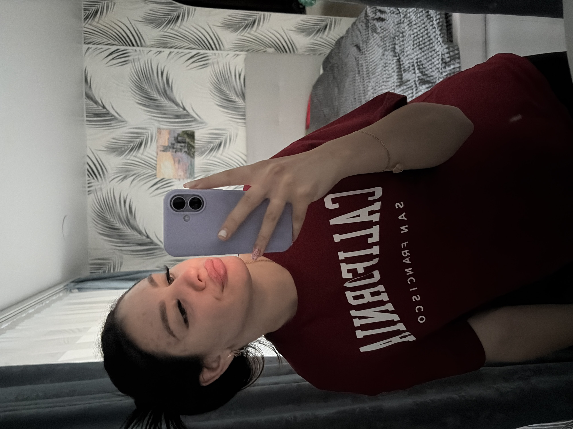 A young woman with dark hair pulled back into a messy bun takes a mirror selfie in a room. She is wearing a dark red t-shirt with white lettering that reads "CALIFORNIA" and "SAN FRANCISCO" in an arc. Her lips are pursed, and she is holding a light purple smartphone in her right hand. The background shows a wall with a palm leaf wallpaper pattern, a small painting of a castle, and a bed with a gray comforter. A dark gray curtain is visible on the left side of the image. The location is not identifiable as Fokino, Russia.