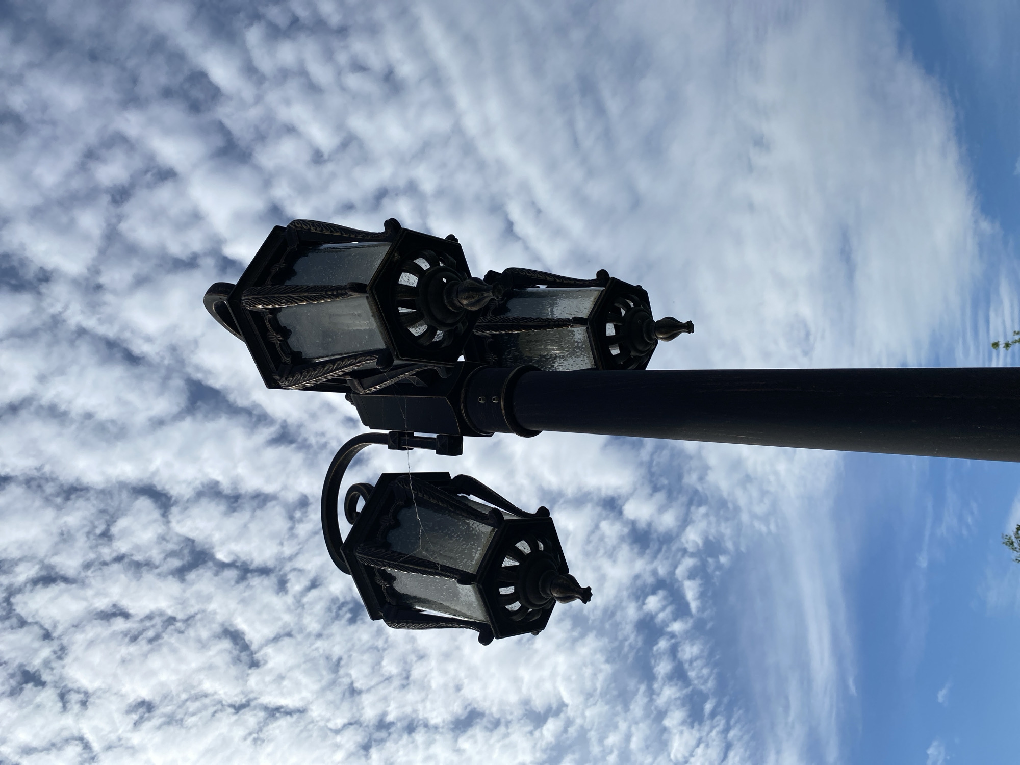 An ornate, dark metal street lamp with three hexagonal lanterns is captured against a bright sky in an upward-angled shot. The image appears rotated, with the lamp pole extending horizontally from the right side of the frame, and the lanterns hanging downwards from it. Each lantern is crafted from dark, possibly wrought iron or cast metal, featuring decorative twisted rope patterns and pointed finials at the bottom. The glass panels of the lanterns are textured or frosted, diffusing the light. Fine cobwebs are visible on one of the lanterns, suggesting it has been exposed to the elements.
The background is a dynamic sky, transitioning from dense, altocumulus-like clouds with distinct patterns on the left to more wispy, cirrus-like streaks on the right, where patches of clear blue sky are visible. The interplay of light and shadow highlights the silhouette of the lamp against the bright, cloudy backdrop, suggesting it is daytime. No people or text are visible. The setting is an outdoor public space in Tyumen, Russia.