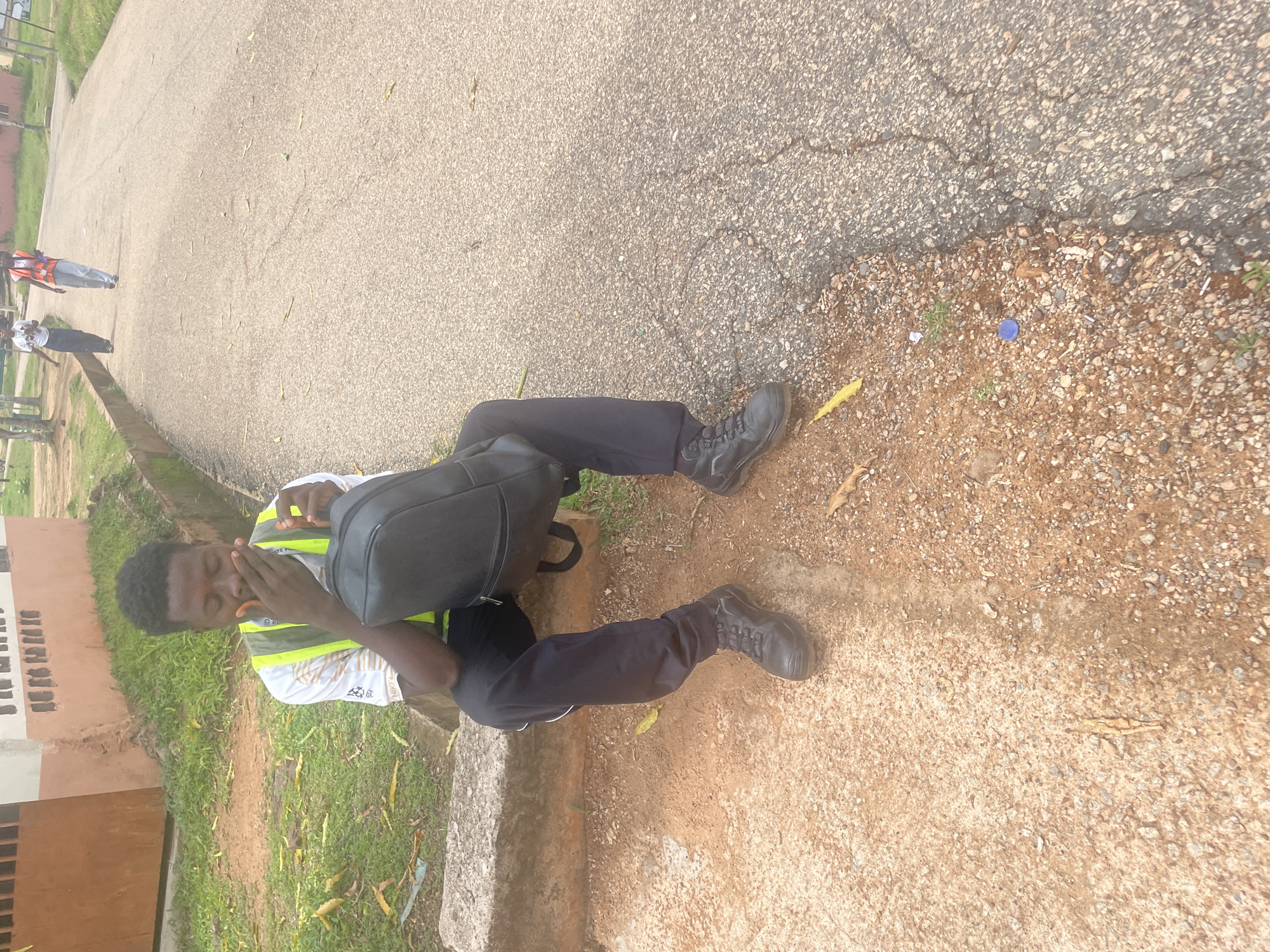 A young Black male wearing a white shirt with a reflective vest over it, dark pants, and black boots is sitting on a concrete curb. He is holding a black backpack on his lap and covering his mouth with his hand, appearing to yawn or cough. The background shows a paved road with two individuals walking away in the distance, also wearing reflective vests. To the left of the man is a patch of grass and a building with brown and white walls. The scene appears to be outdoors, possibly on a street or walkway in Lokoja, Nigeria, given the building style and vegetation. The ground is a mixture of dirt, gravel, and asphalt.