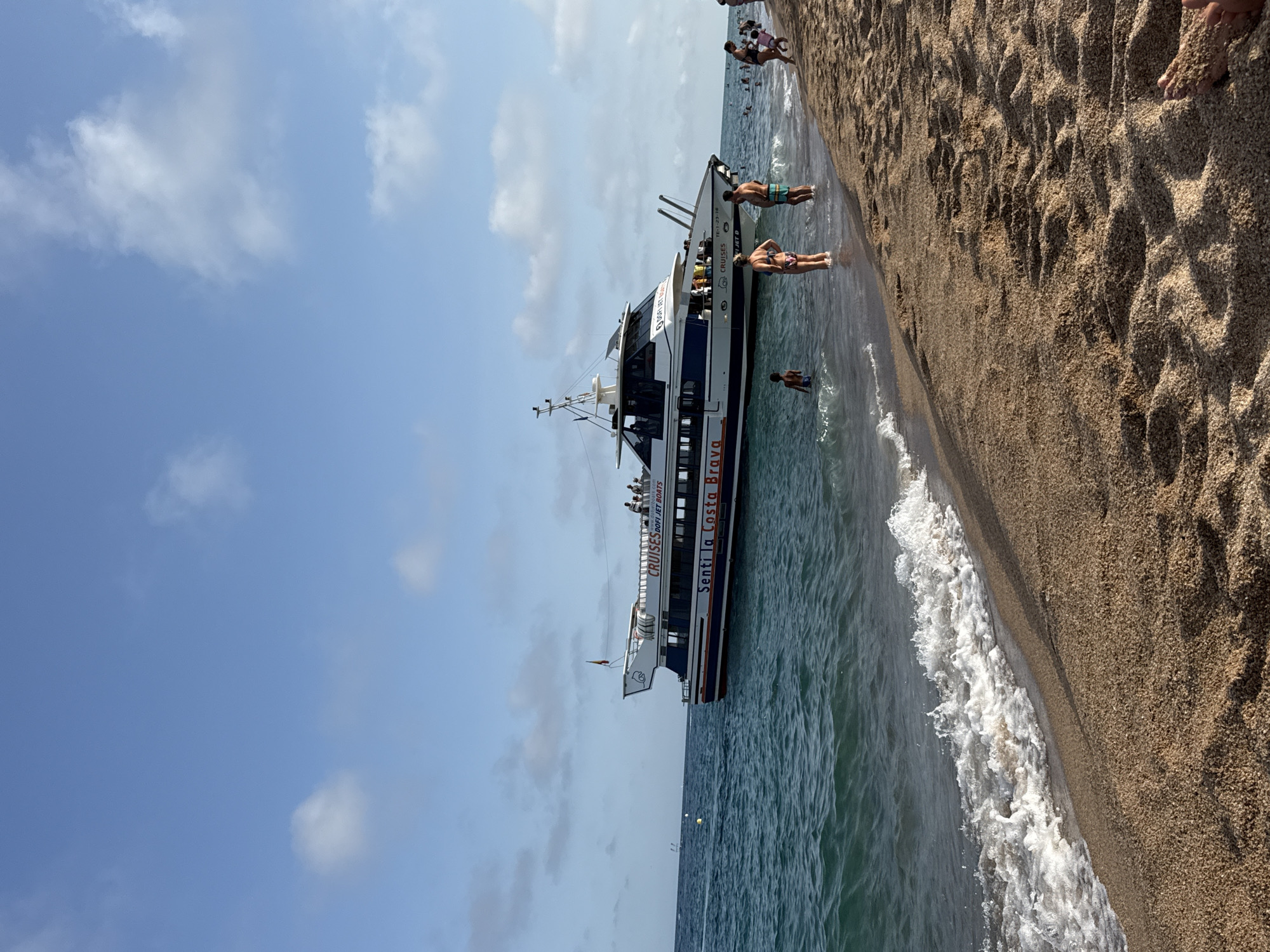 In Blanes, Spain, on a sandy beach with gentle waves lapping the shore, a large white and blue passenger boat named "Senti la Costa Brava" is anchored offshore. The boat, which appears to be a tourist cruise vessel, has a dark blue stripe along its hull and the words "CRUISES DOFI JET BOATS" printed on its side. A Spanish flag flies from the top deck. Several people are visible on the boat's upper deck, and a few individuals are standing in the shallow water near the shore. On the beach, more people are present, some wading in the water and others walking along the sandy expanse. The sky above is a clear blue with scattered white clouds, indicating fair weather. The scene captures a typical summer day at the beach, with recreational activities taking place both on the water and on land.