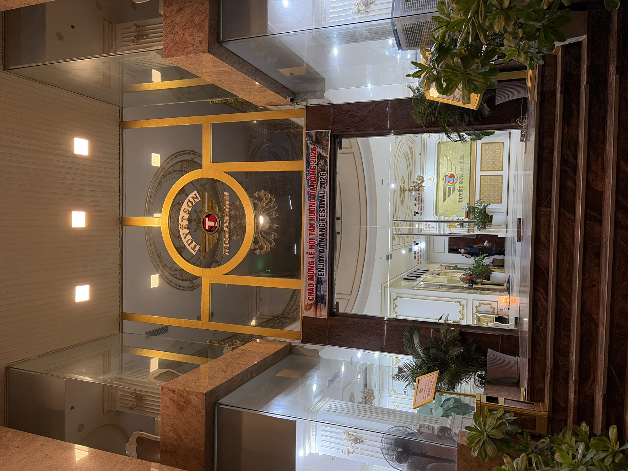 The image shows the entrance to the Tuyet Son Hotel in Da Nang, Vietnam. The hotel has a grand entrance with marble columns, gold trim, and a large glass facade. Above the entrance, a circular logo with the hotel's name is visible. A banner across the entrance reads "CHAO MUNG LE HOI TAN HUONG DA NANG 2026" and "ENJOY DA NANG FESTIVAL 2026". Inside the hotel, a reception desk with the hotel's logo is visible, and a few people are seen in the lobby. Plants and decorative elements are placed throughout the entrance area. The lighting suggests it is either dusk or nighttime, with artificial lights illuminating the scene.