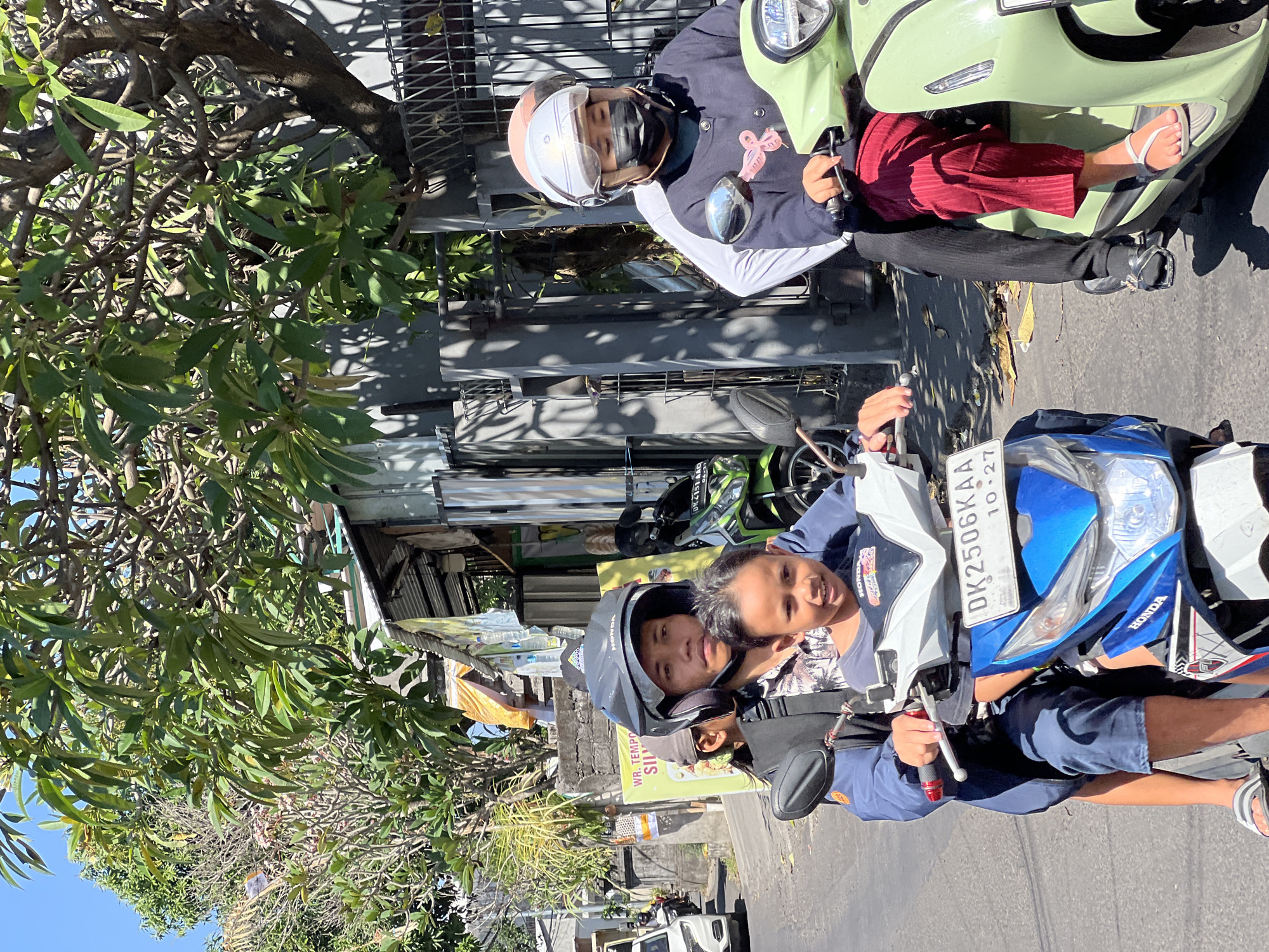 Two motorbikes are traveling on a street in Denpasar, Indonesia. On the first motorbike, a young boy sits in front of an adult male who is driving. Both are wearing helmets. The boy is smiling at the camera. The motorbike is blue and white. On the second motorbike, a female is driving, and behind her is another person. The female is wearing a helmet and a face mask. The motorbike is light green. The street is paved, and there are buildings and trees on either side. The sky is clear and blue.