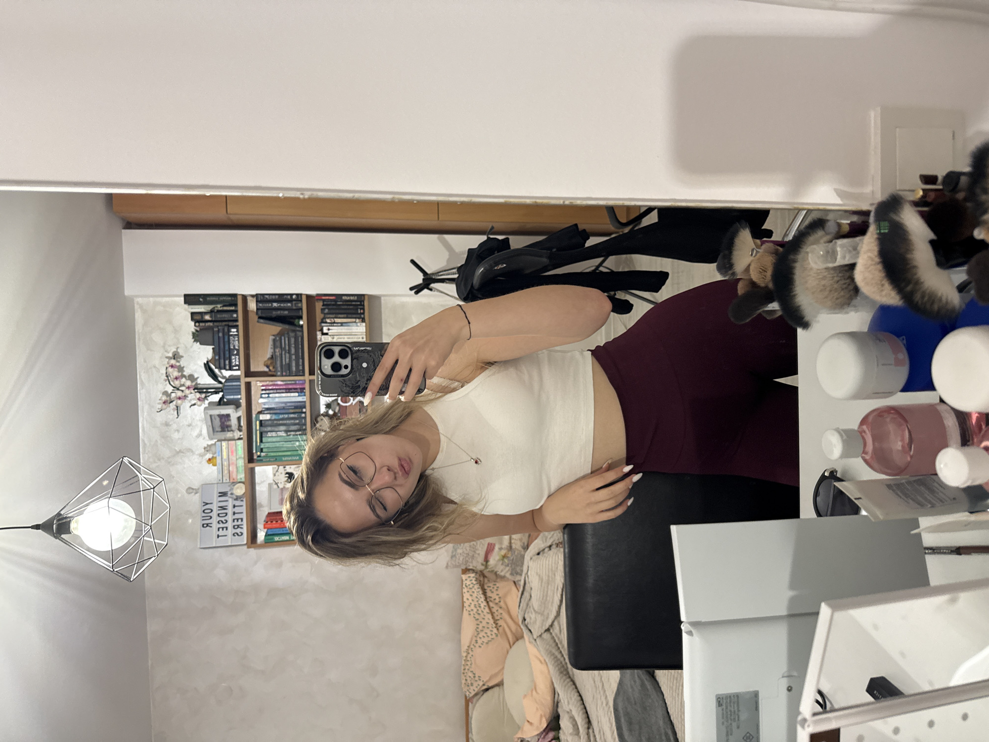 A young woman wearing glasses, a white top, and maroon leggings is taking a selfie in front of a mirror. The mirror reflects a room with a bookshelf, a hanging light fixture, and a sign that reads "YOUR MINDSET MATTERS". A makeup desk cluttered with brushes and various cosmetic products is positioned to the right of the mirror. The location is likely a residential interior in Blagoevgrad, Bulgaria.