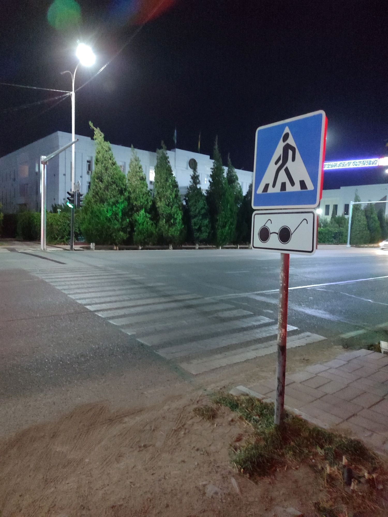 A nighttime street scene in Nukus, Uzbekistan, features a pedestrian crossing and a traffic sign. A prominent blue triangular sign with a white pedestrian silhouette indicates a crosswalk. Below it, a rectangular sign displays a pair of black-rimmed glasses. The signs are mounted on a metal pole with red and gray markings. A pedestrian crossing, marked by white stripes, stretches across the asphalt road. In the background, a modern building with illuminated windows stands amidst lush green trees. A streetlamp casts a bright light on the scene. The sky is dark, with streaks of light suggesting light pollution or lens flare. The ground is paved with asphalt and sections of concrete tiles, with patches of dirt and grass.