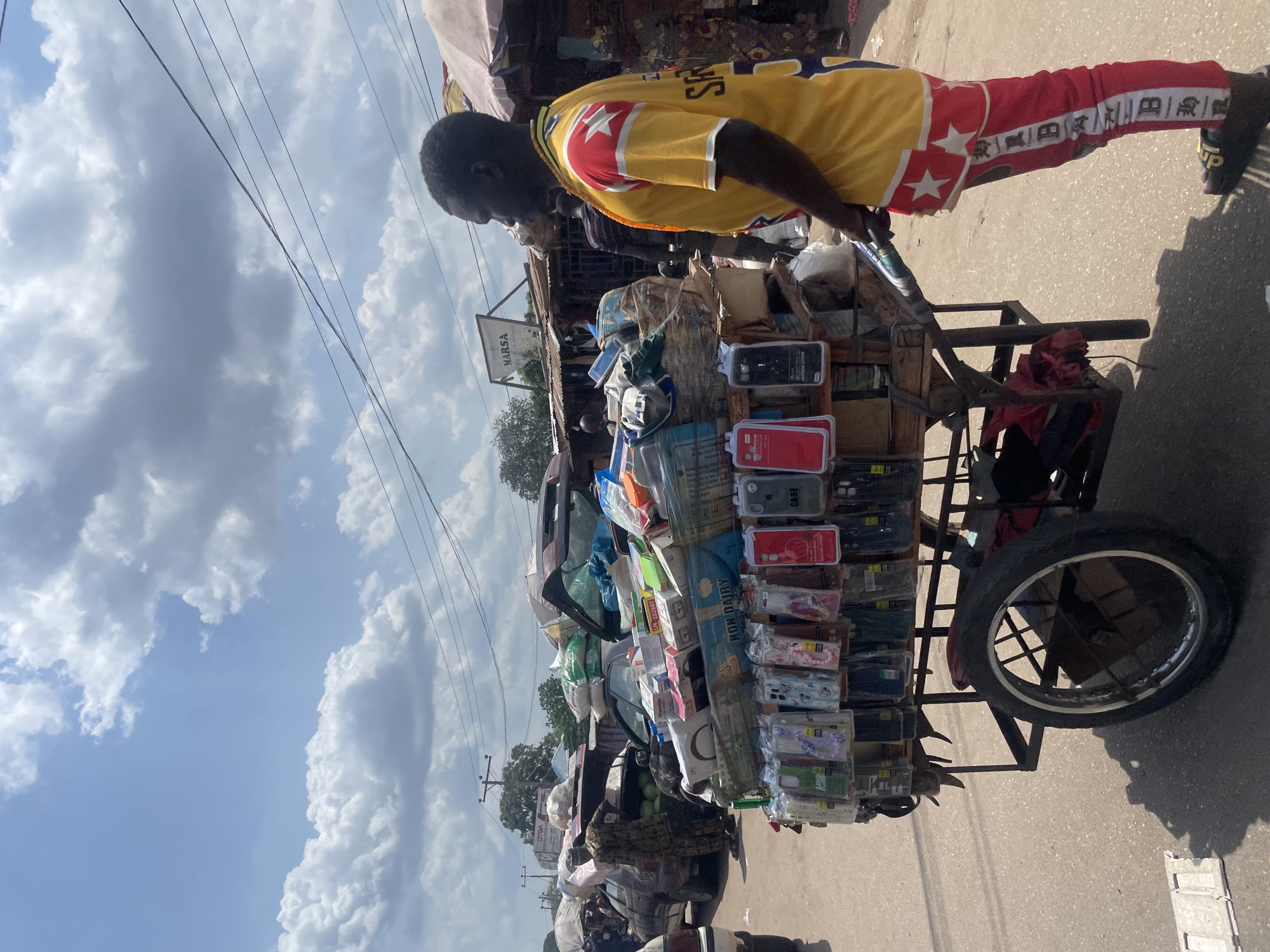 A man pushes a cart loaded with various items in Lokoja, Nigeria. The cart's contents include packaged goods, possibly phone accessories and non-dairy products. The man is wearing a yellow jersey with red accents and patterned red pants. The road is paved, and the sky is visible with scattered clouds. Buildings and electrical lines are visible in the background. The scene is typical of a street market or vendor setup in Lokoja.