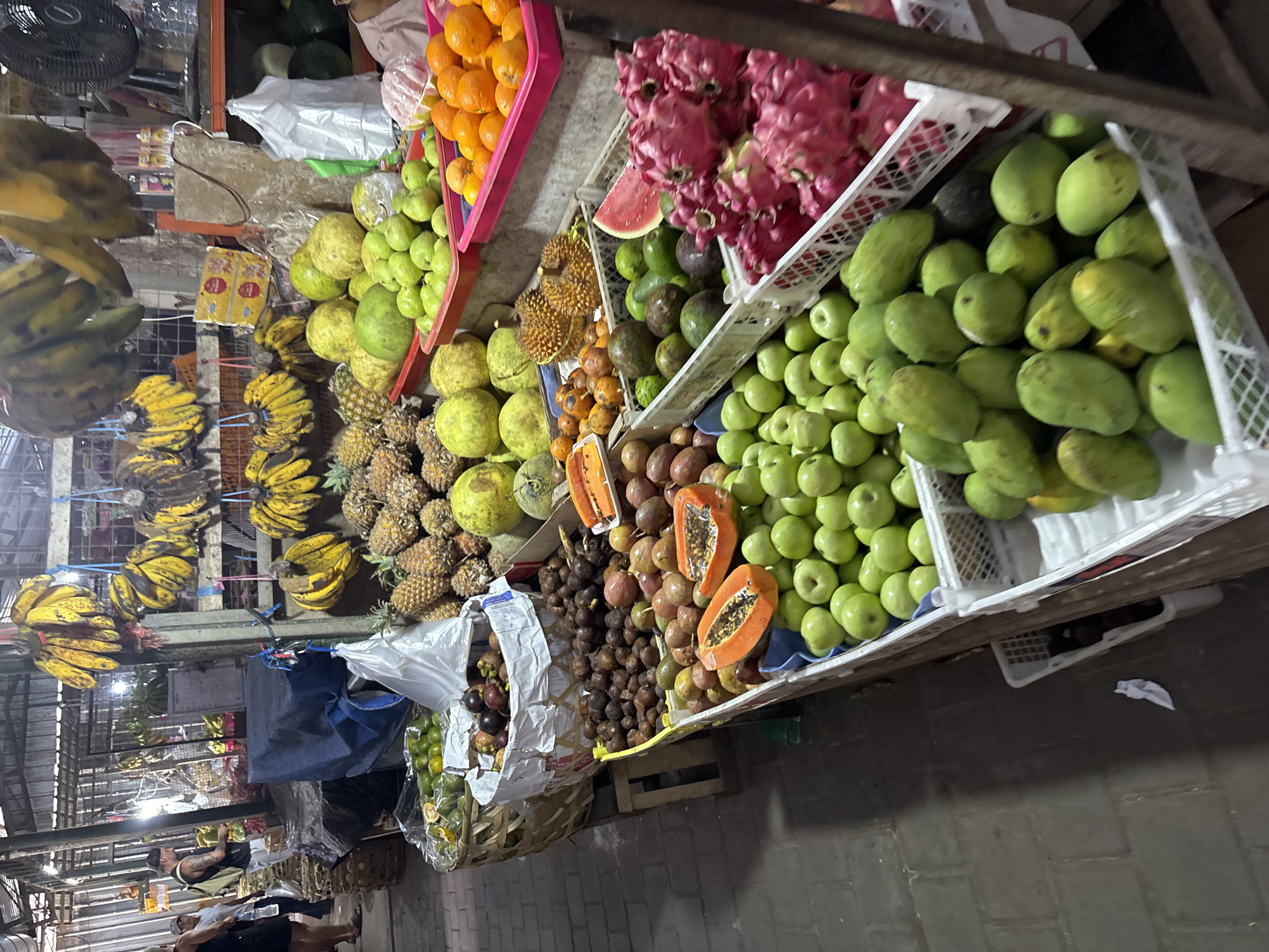 The image shows a fruit stand in Ungasan, Indonesia. The fruit stand displays a variety of fruits, including bananas, oranges, dragon fruit, avocados, apples, mangos, durian, papaya, pineapples, and passion fruit. The fruits are arranged in baskets and crates. Several people can be seen walking past the fruit stand in the background. The setting suggests an outdoor market or street vending area.