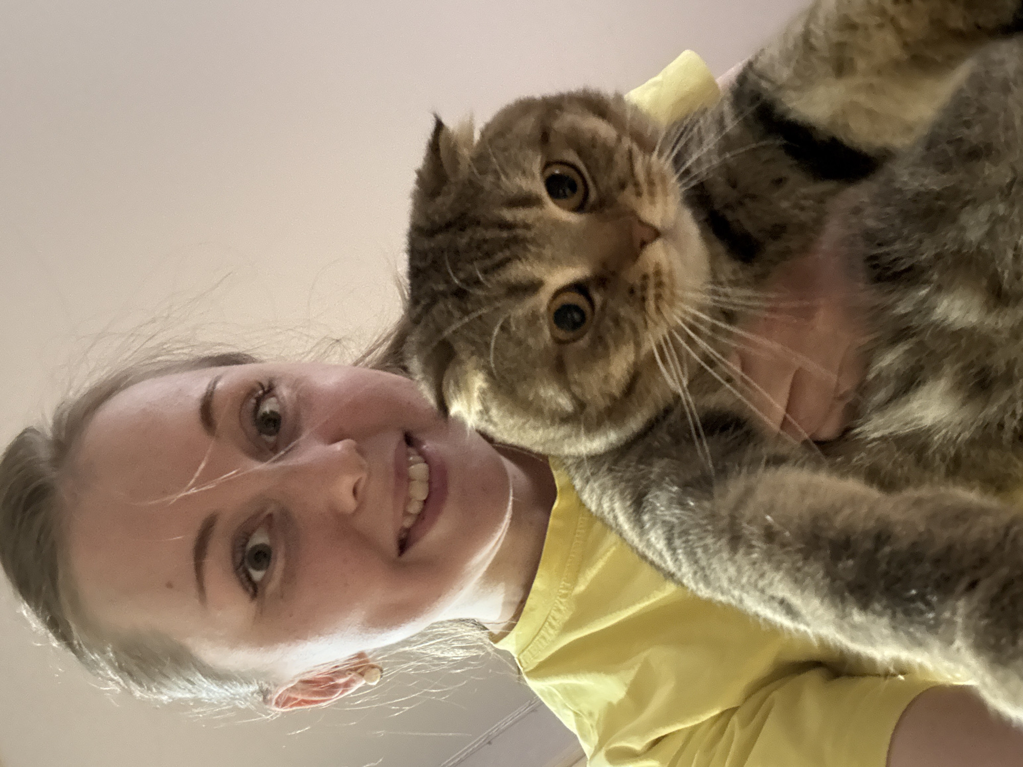 A woman in a yellow shirt holds a brown and gray tabby cat. The woman is smiling at the camera, and the cat is looking at the camera with wide eyes. The background is a plain ceiling. Location: Unknown, Unknown.